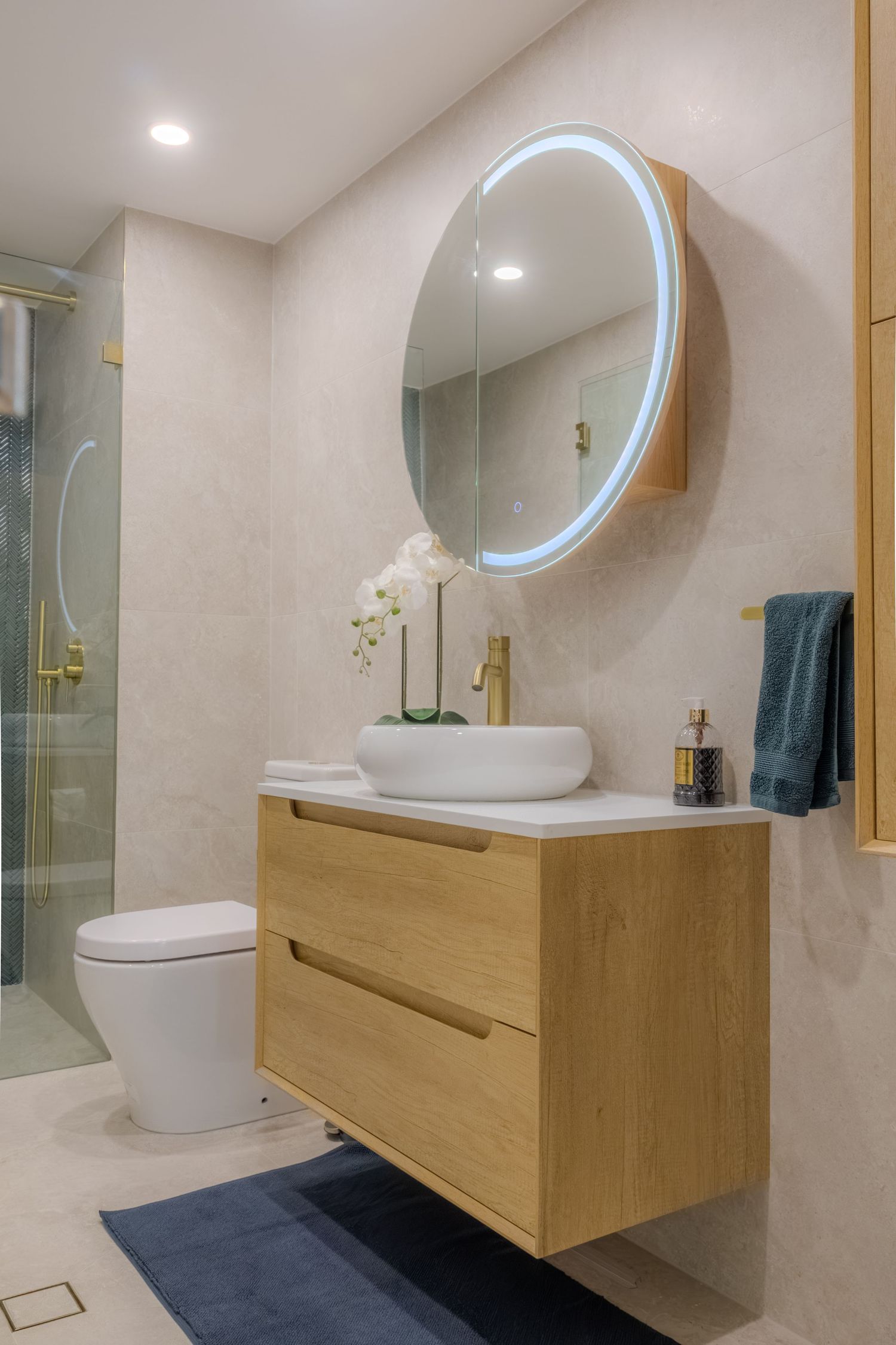 Modern bathroom with wooden vanity, round LED mirror, vessel sink, gold fixtures, and glass shower enclosure.