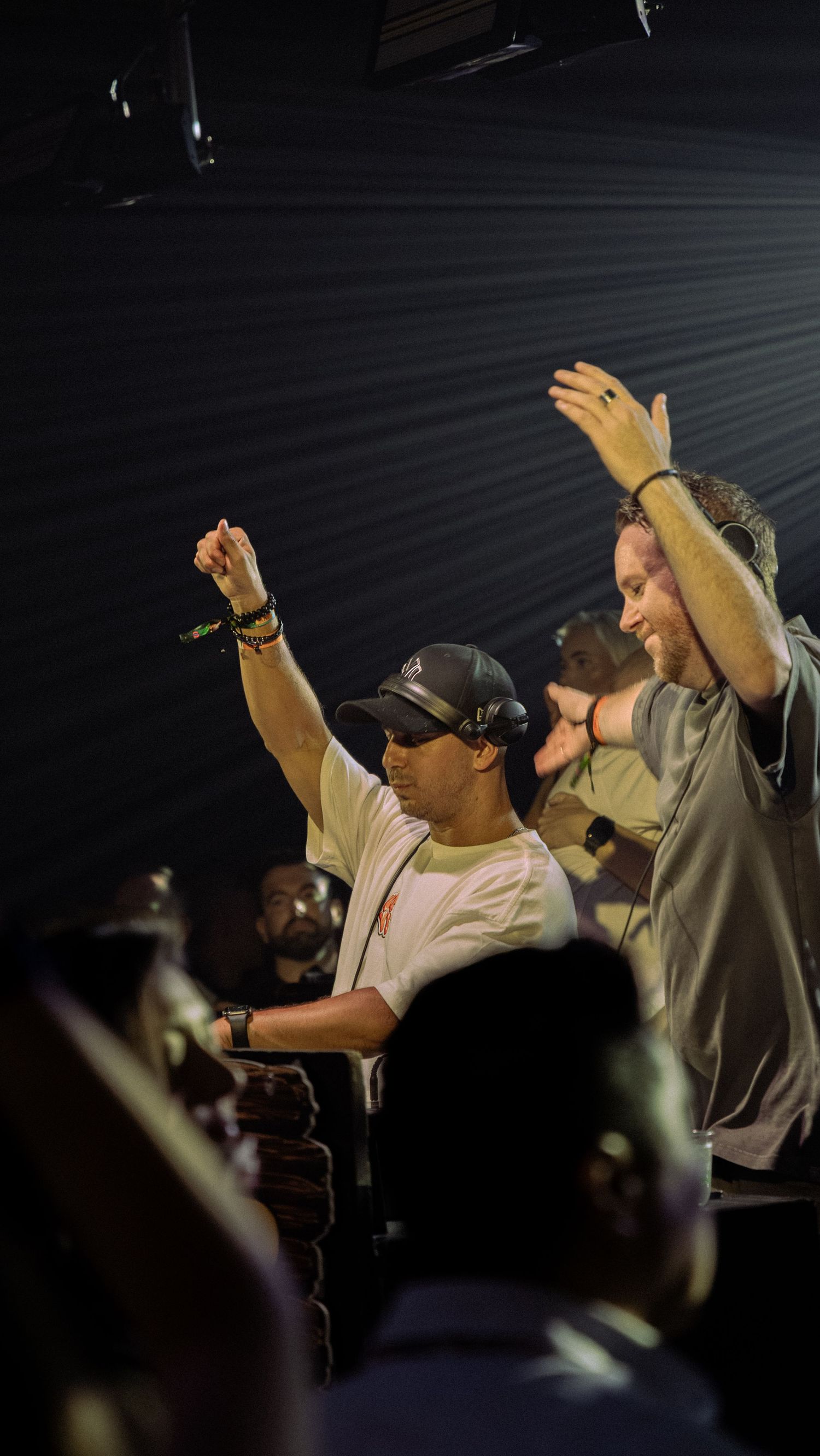 Two men celebrate with raised arms at a lively nightclub event, one wearing a cap and white tee.