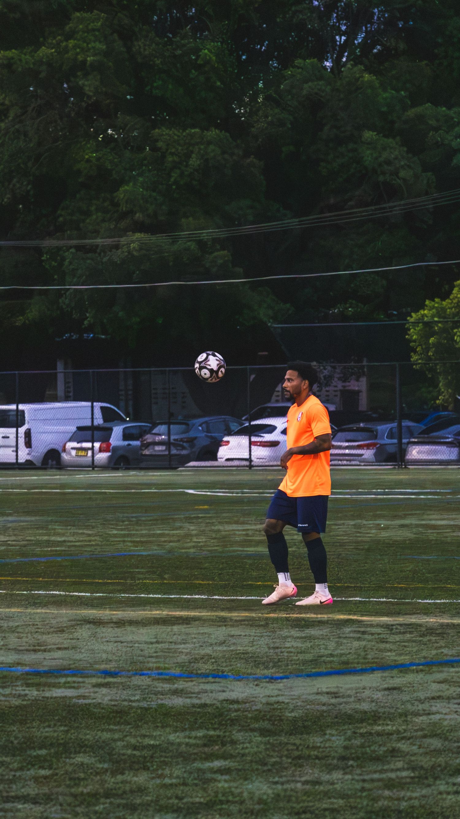 Two soccer players practicing on a grass field with one wearing an orange jersey and a ball in the air.