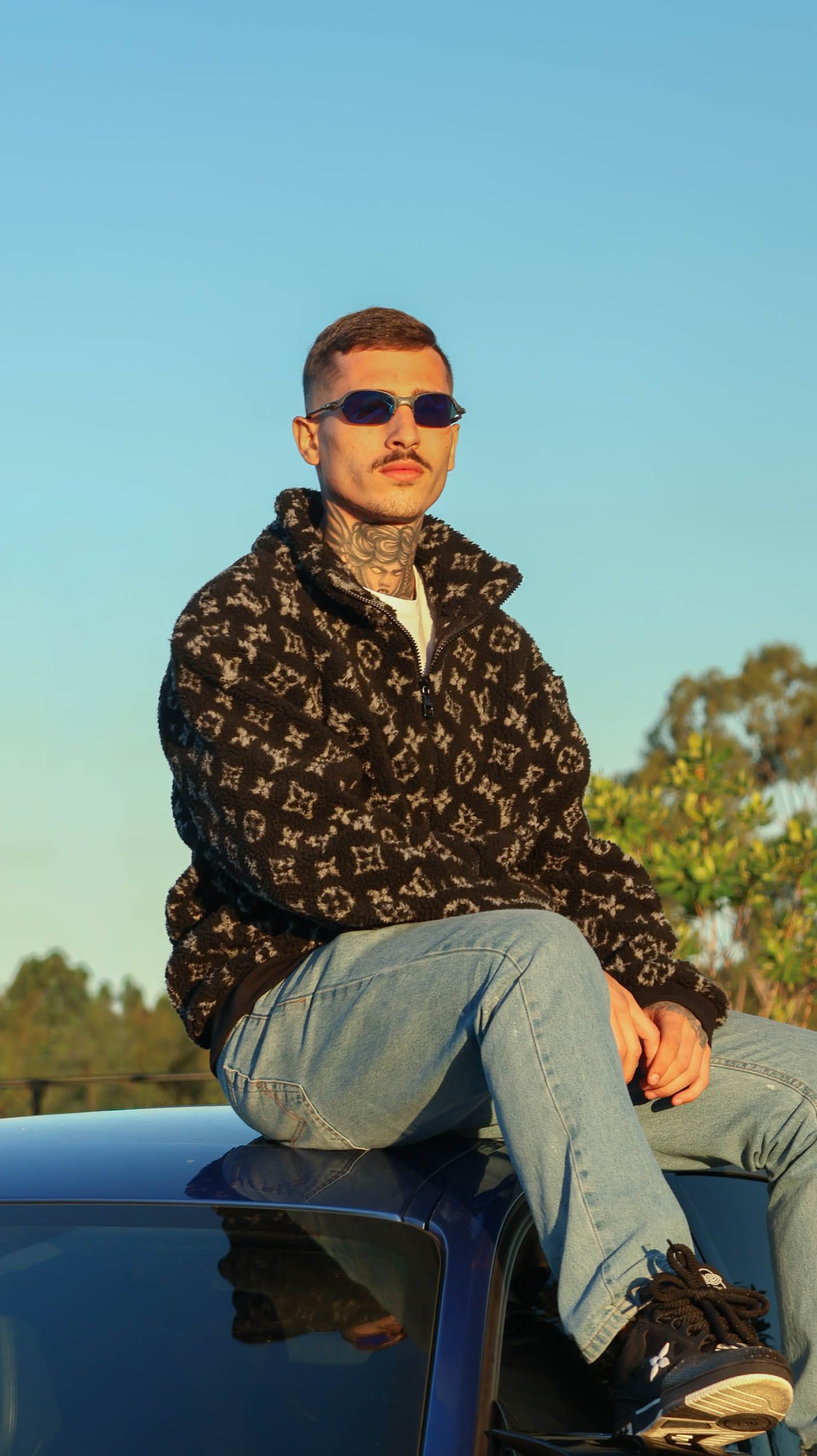 Young man in Louis Vuitton fleece jacket and jeans sits on car hood wearing purple sunglasses outdoors.
