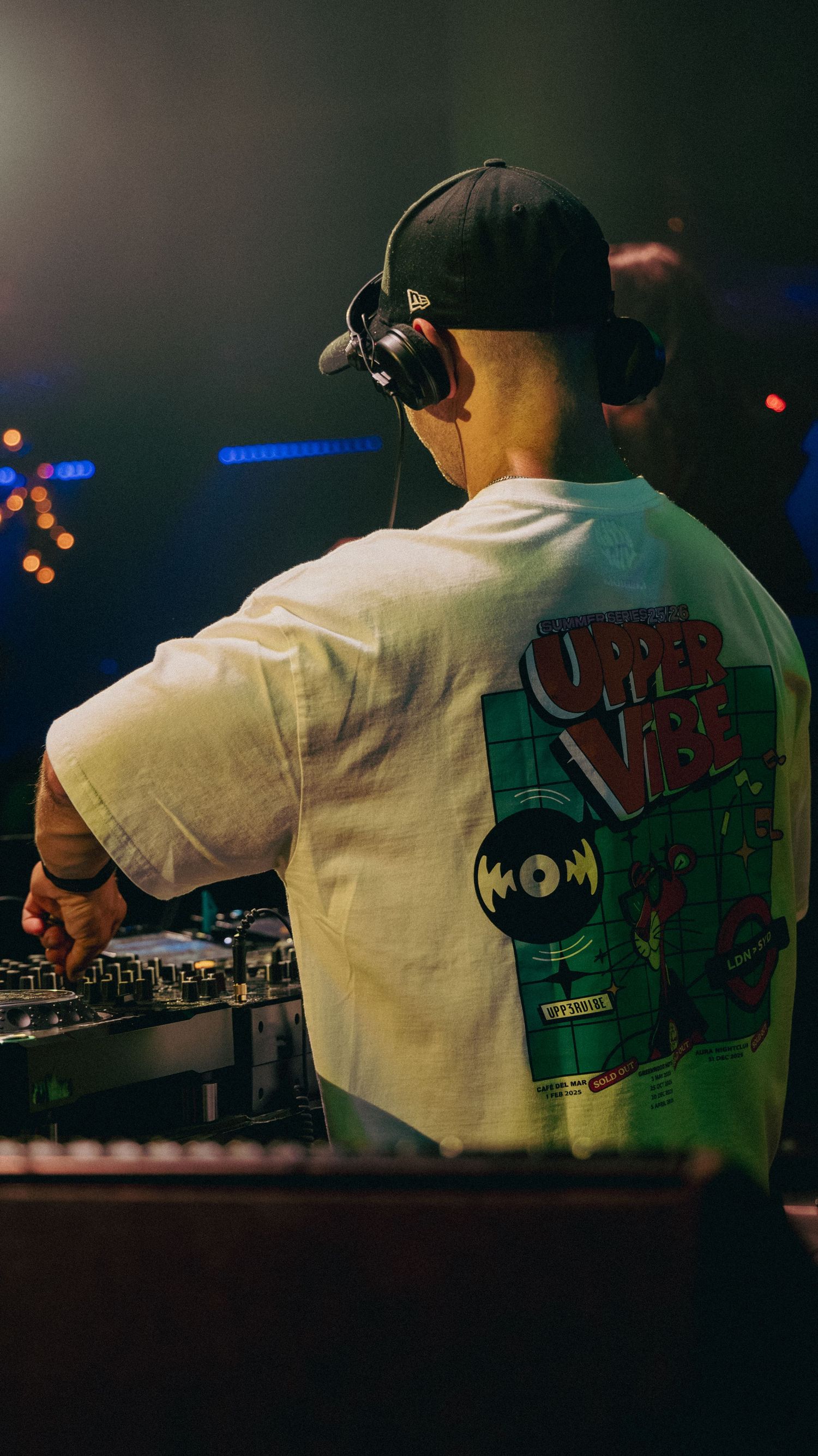 DJ in white graphic tee and cap mixing at turntables in a dark nightclub with colorful bokeh lights.