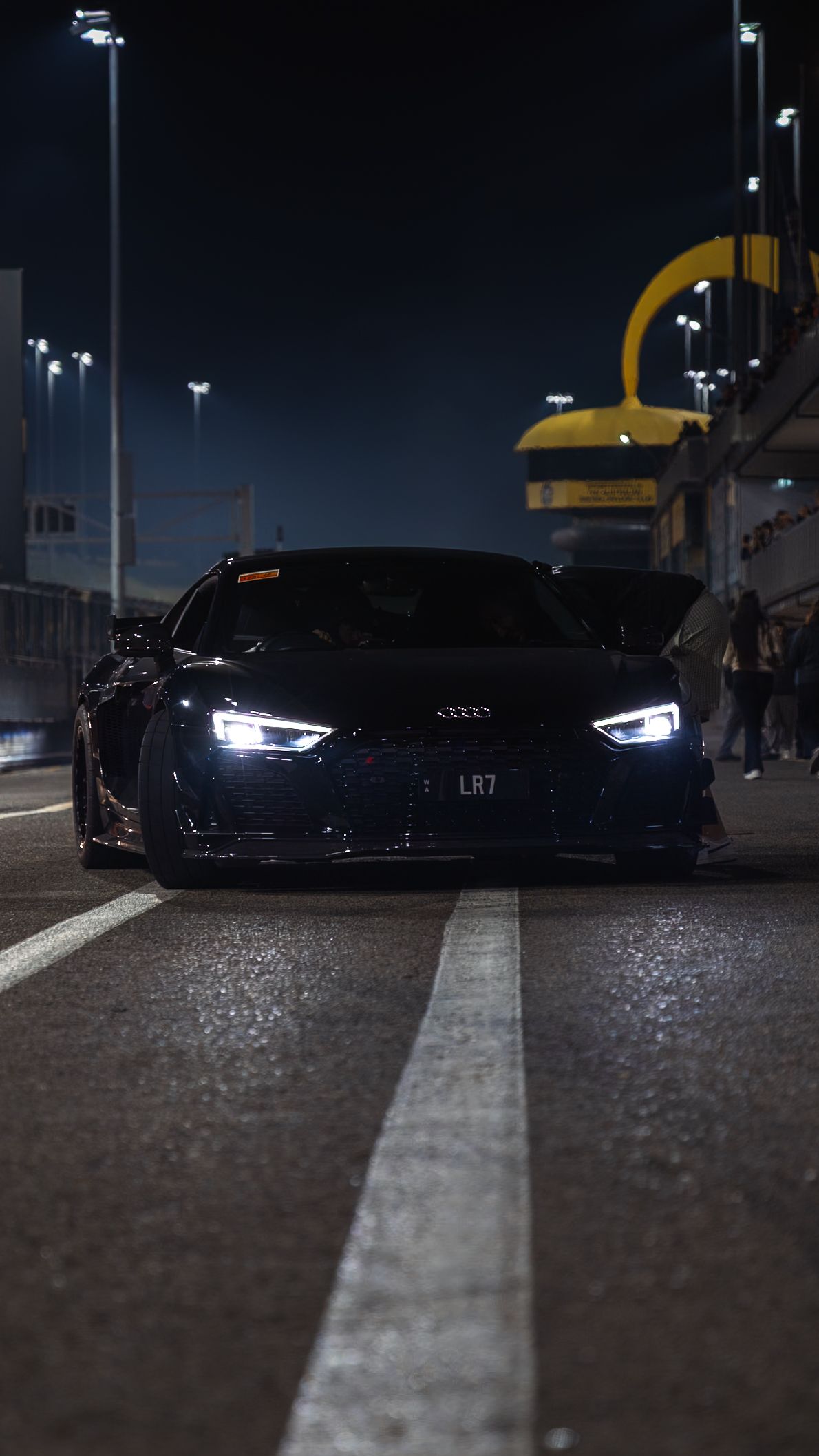 A sleek black Audi R8 with bright LED headlights sits on a wet racing circuit at night.