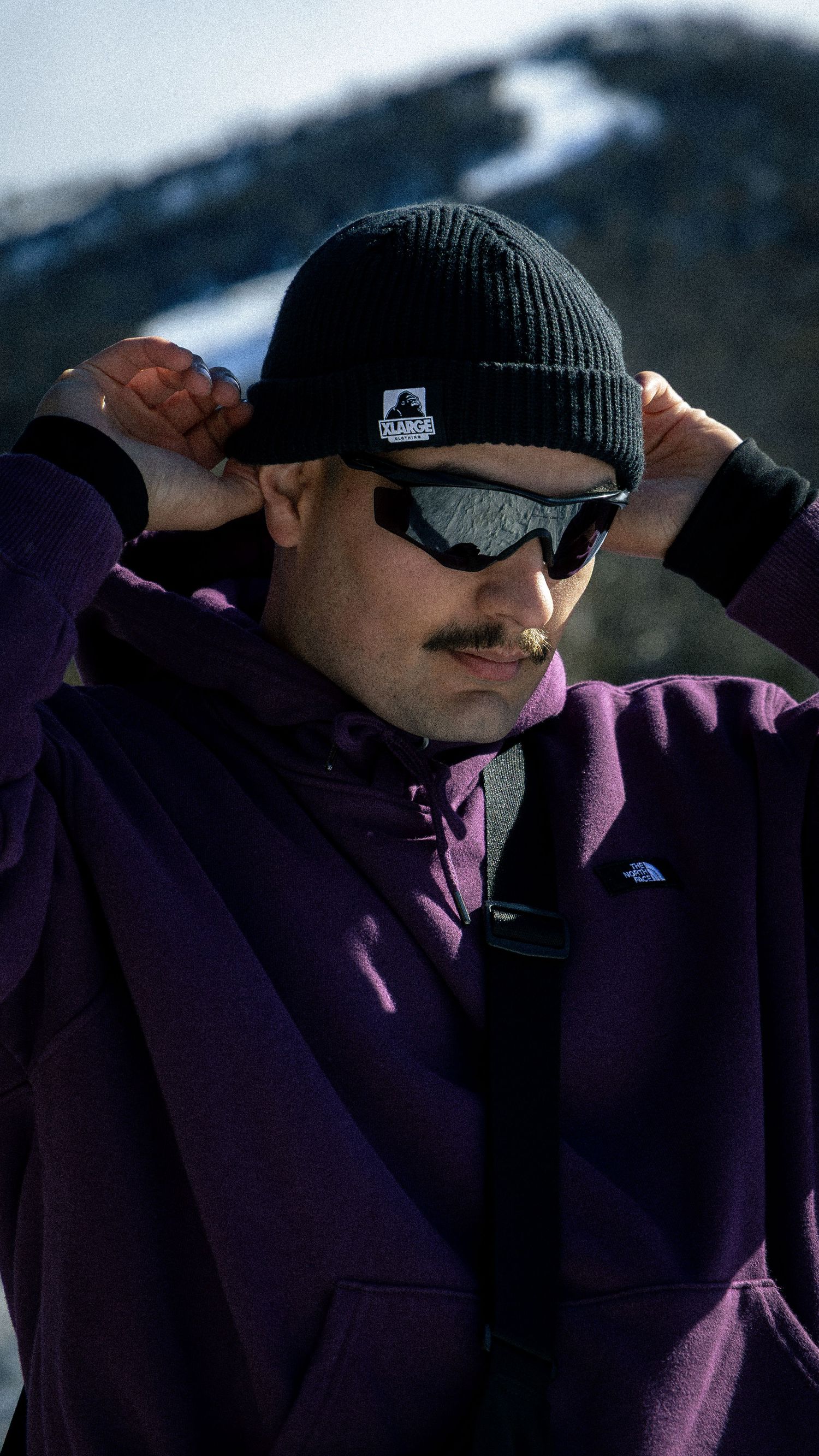 Man in purple jacket and black beanie adjusts hat while wearing sunglasses on a snowy mountain.