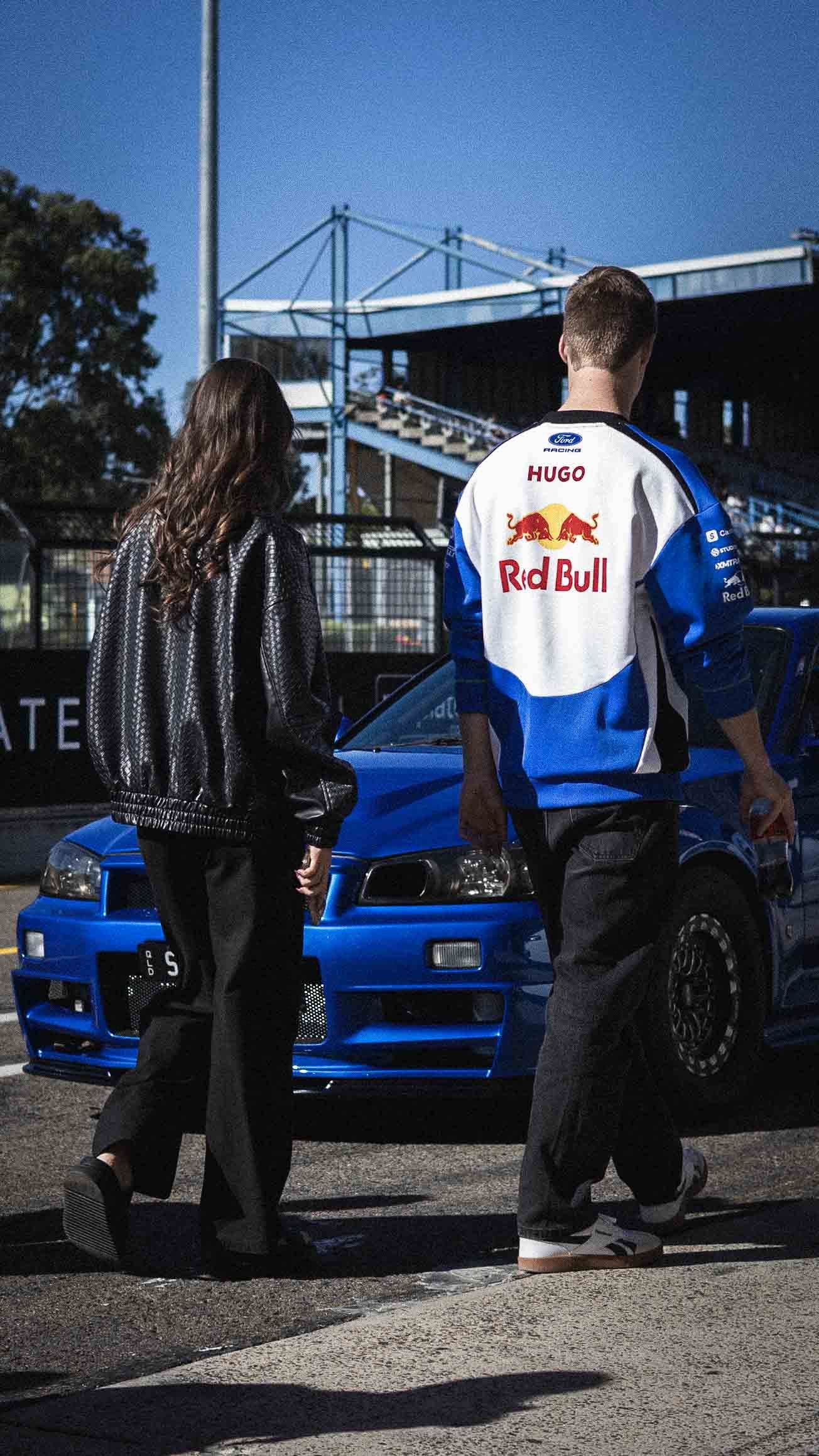 Two people stand before a blue Ford race car, one wearing a Hugo Red Bull racing jersey at an outdoor motorsport venue.