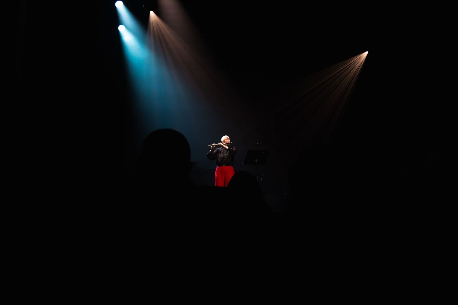 A lone performer in red stands under dramatic stage spotlights in a dark concert venue.
