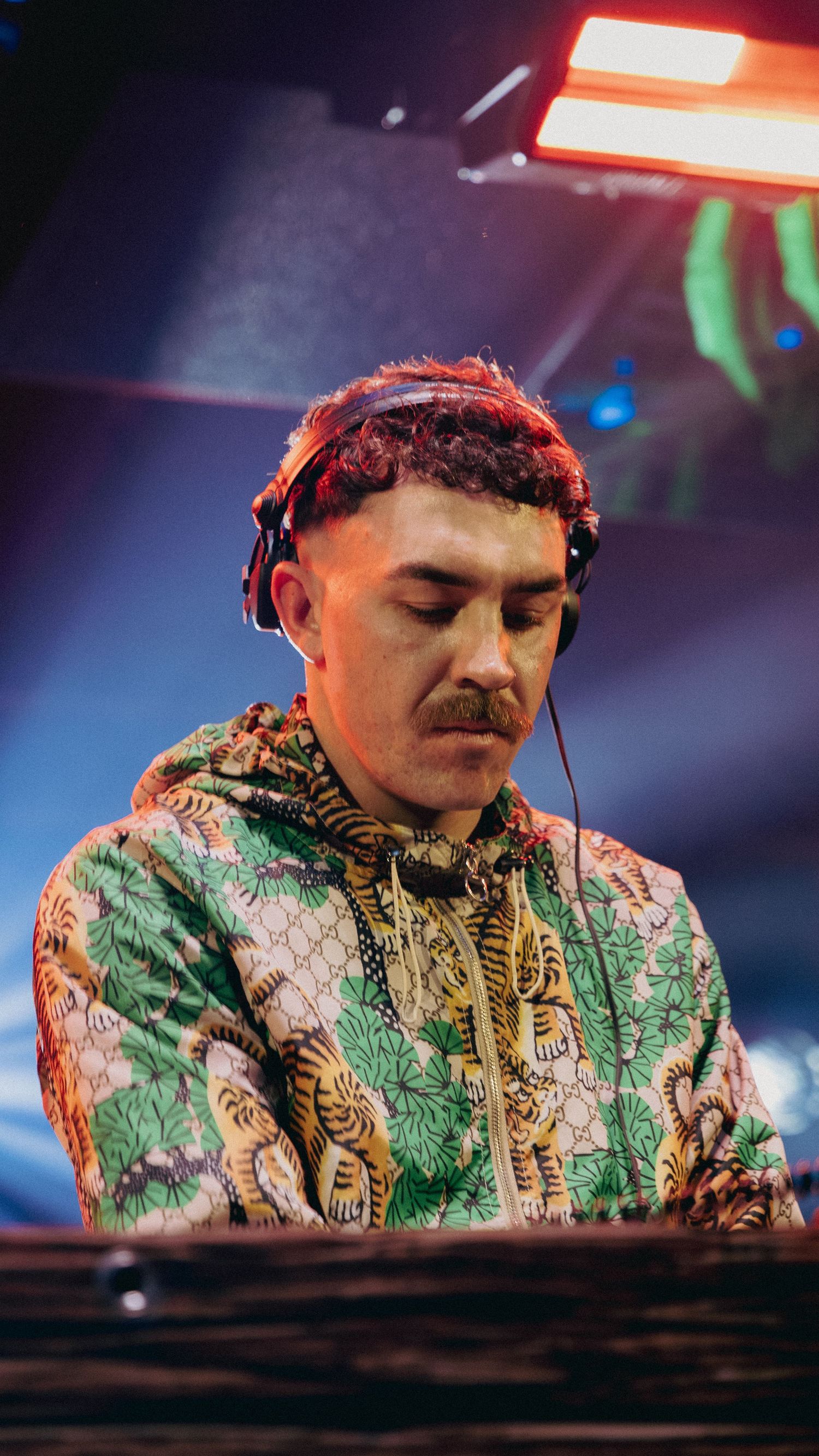 A DJ wearing headphones and a colorful tiger-print hoodie performs at a nightclub under red stage lighting.
