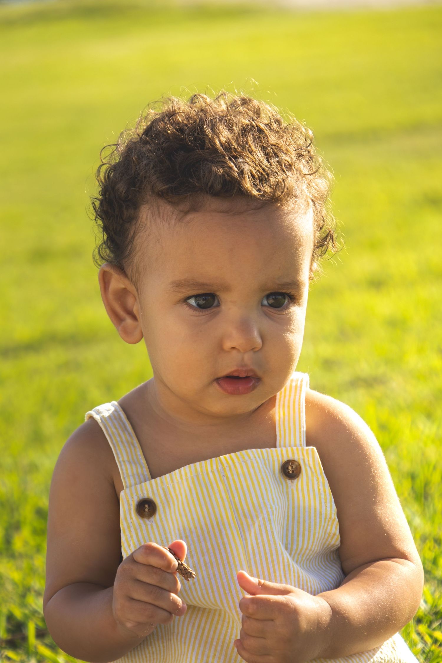 Baby in white tank top outfit plays in sunny green grass field.