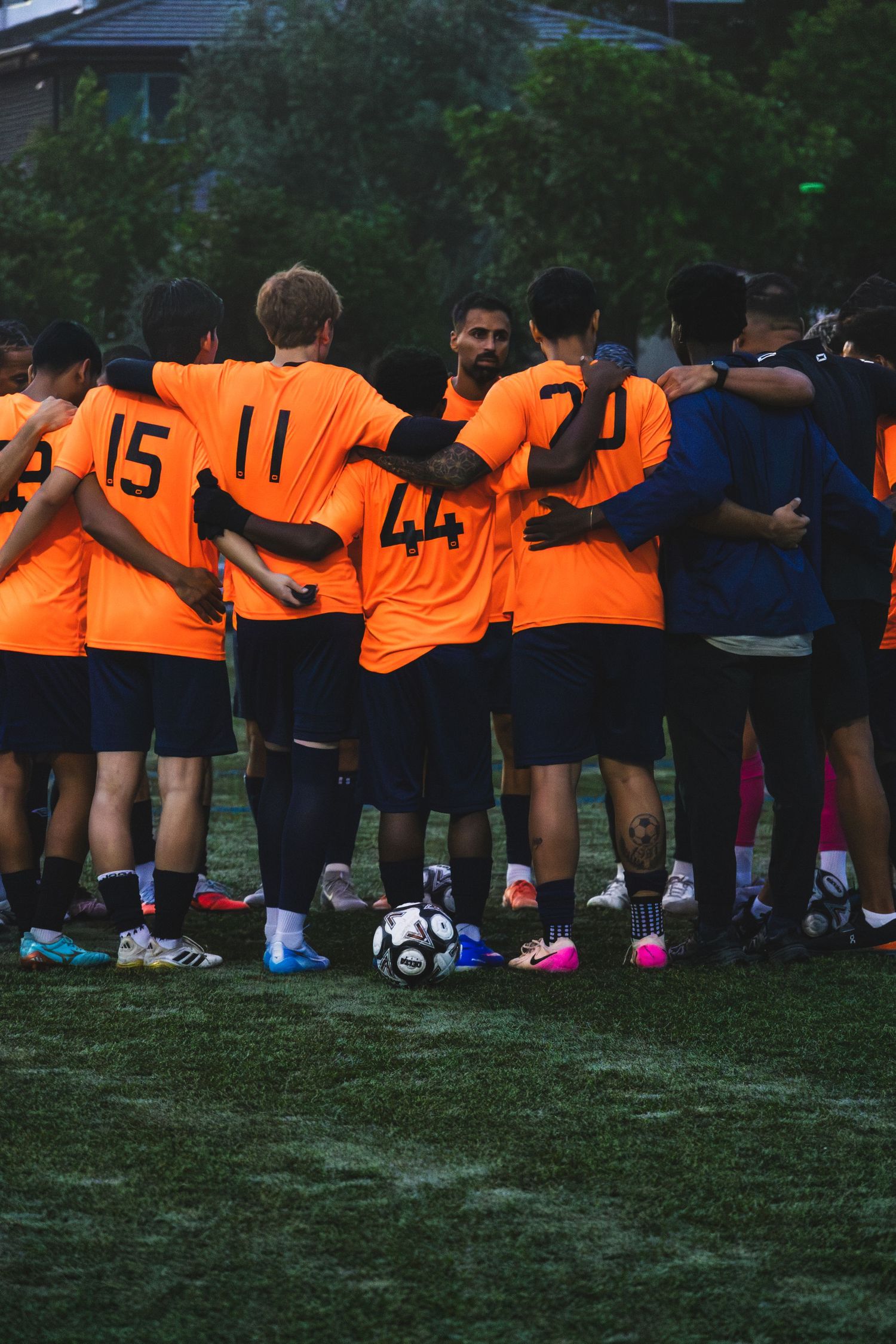 Soccer team in orange jerseys huddles together on field with soccer ball visible at their feet during practice.