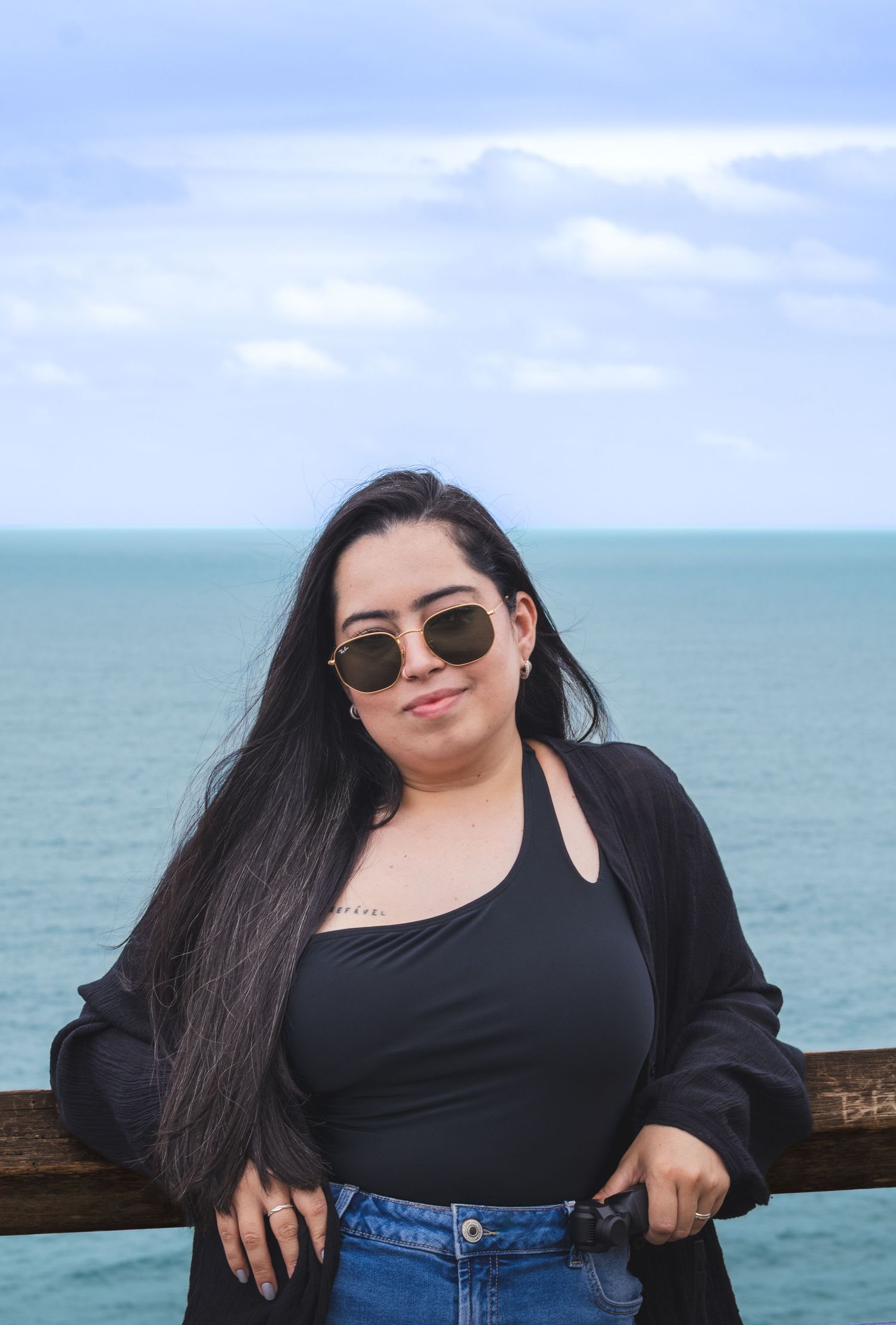 A person in a black top and jeans poses against a railing with the ocean and cloudy sky in the background.