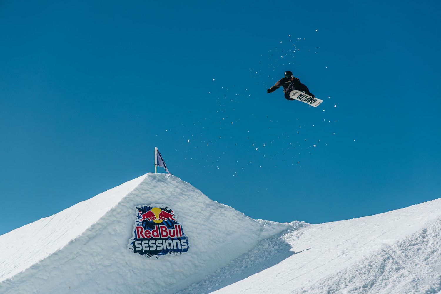 Snowboarder performs a high aerial trick off a snow jump at Red Bull Sessions event against a clear blue sky.