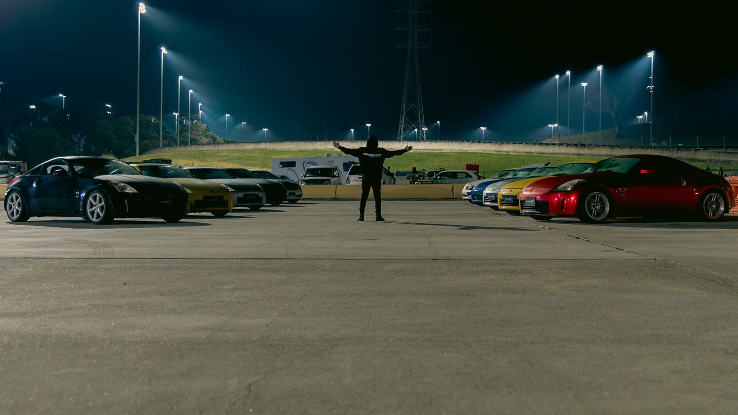 Person standing between two rows of sports cars in a parking lot at night under bright lights.