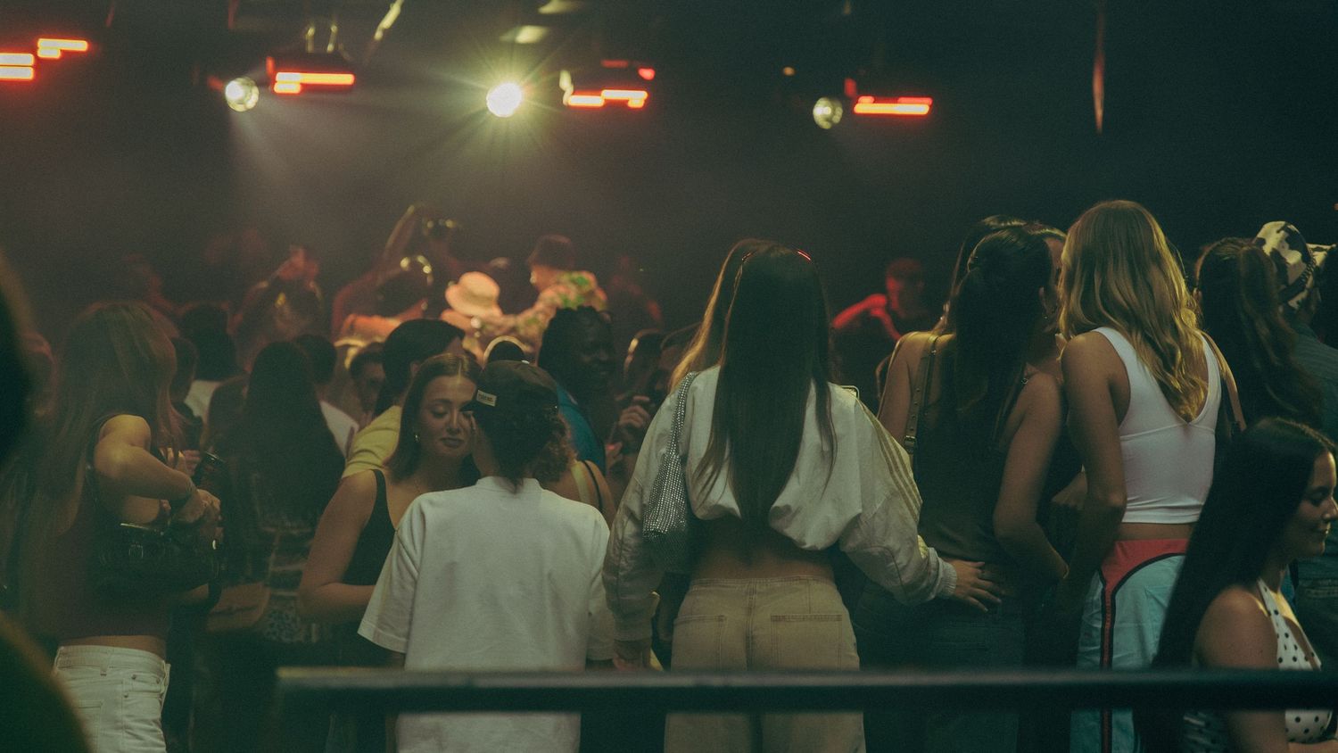 Crowded nightclub dance floor with young people socializing under colorful stage lights in a dark venue.