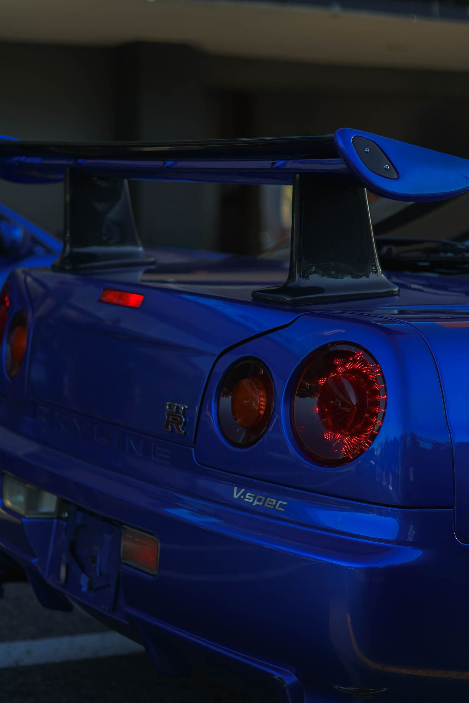 Blue Nissan Skyline GT-R R34 V-Spec rear view with red tail lights and large spoiler in dark parking garage.