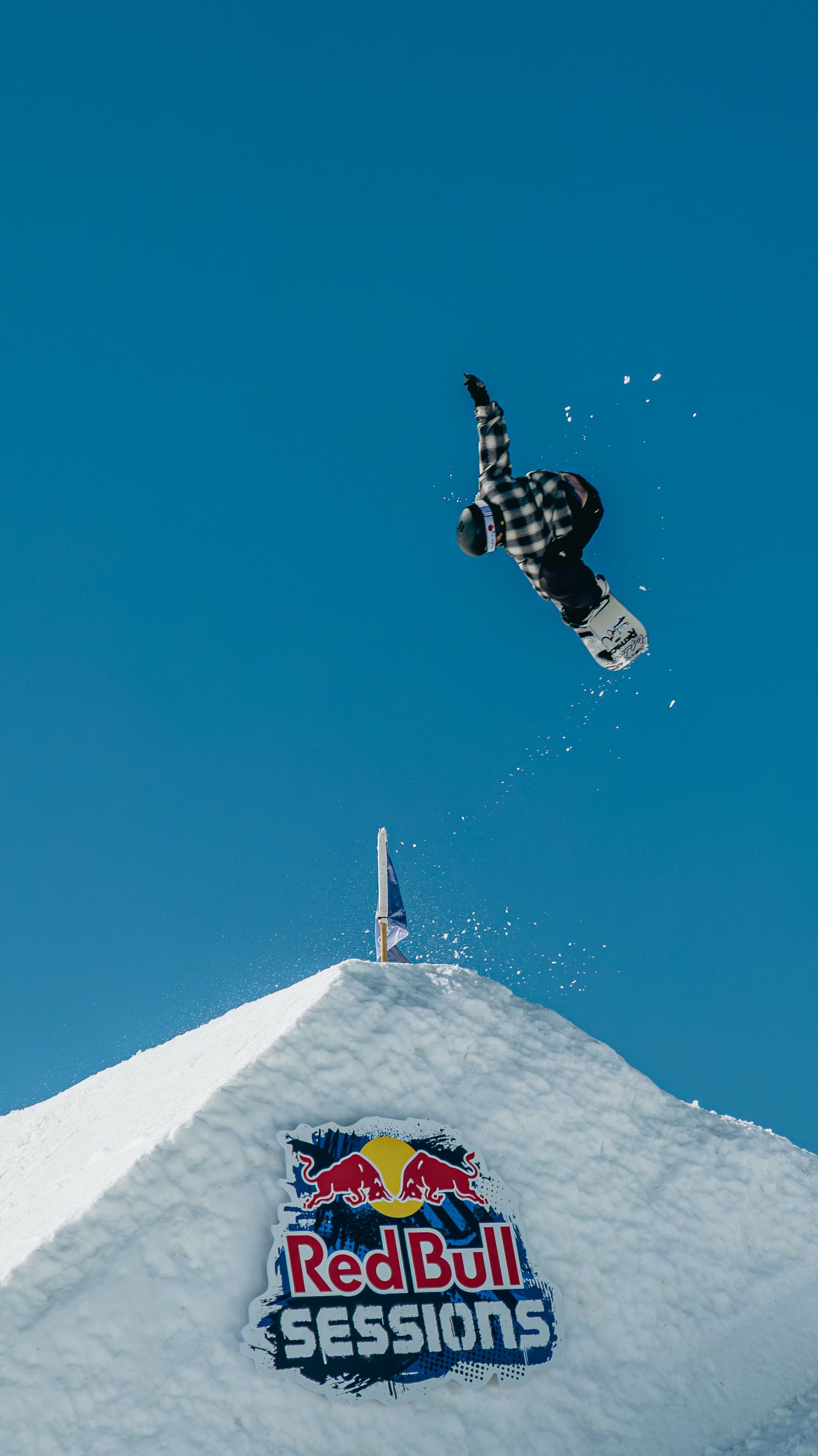 Snowboarder performs aerial trick off a snow ramp at Red Bull Sessions event against a clear blue sky.