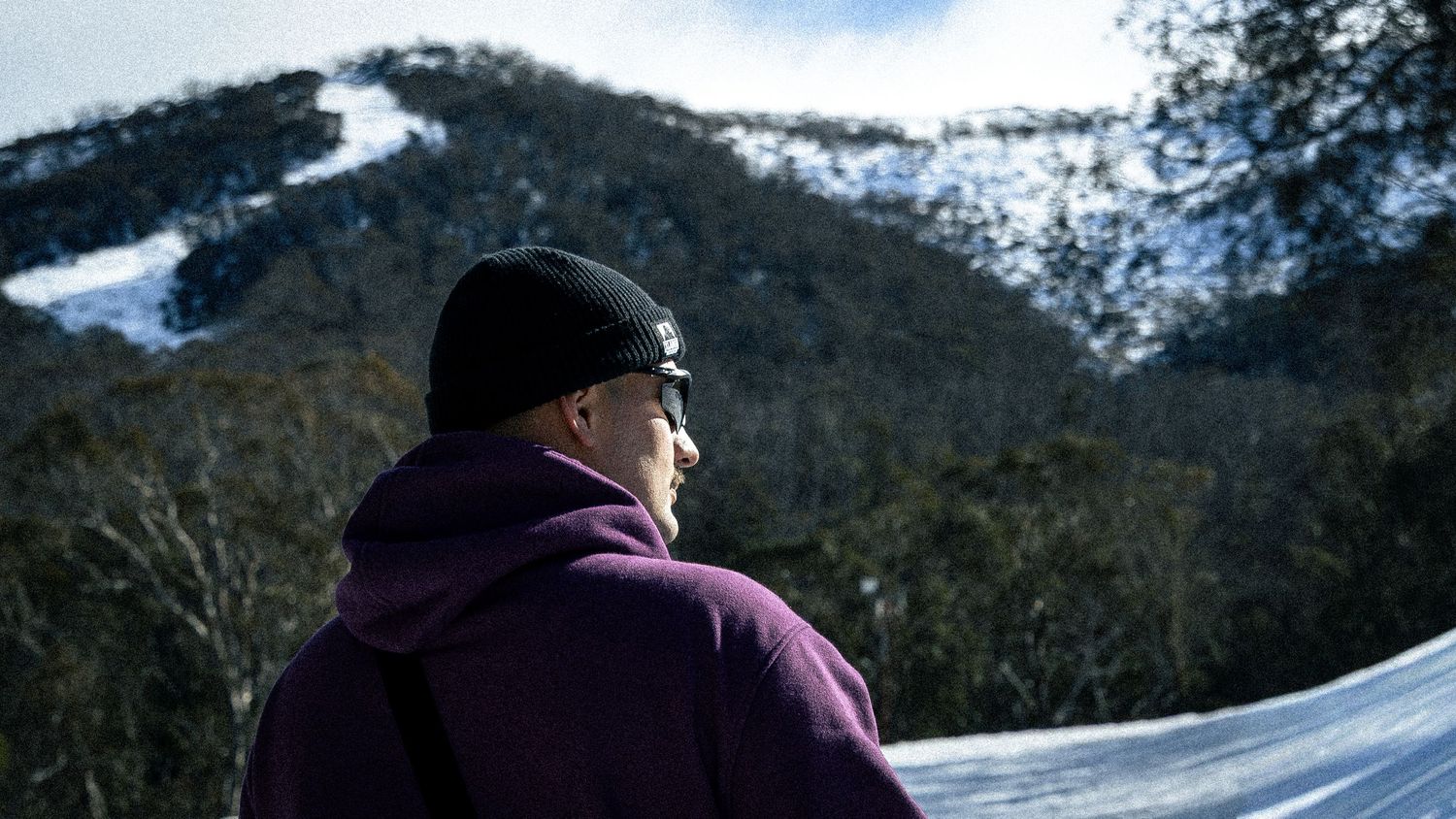 Person in purple hoodie and black beanie gazing at snow-covered mountain landscape on a clear winter day.