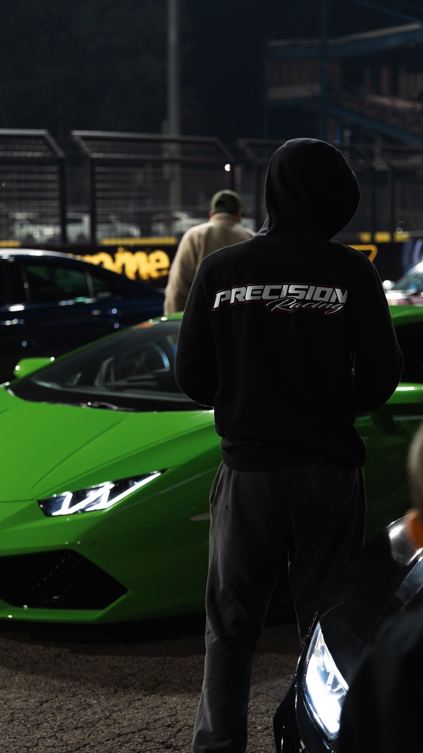 Person in Precision Racing hoodie standing beside a green Lamborghini Huracan at a night car meet.