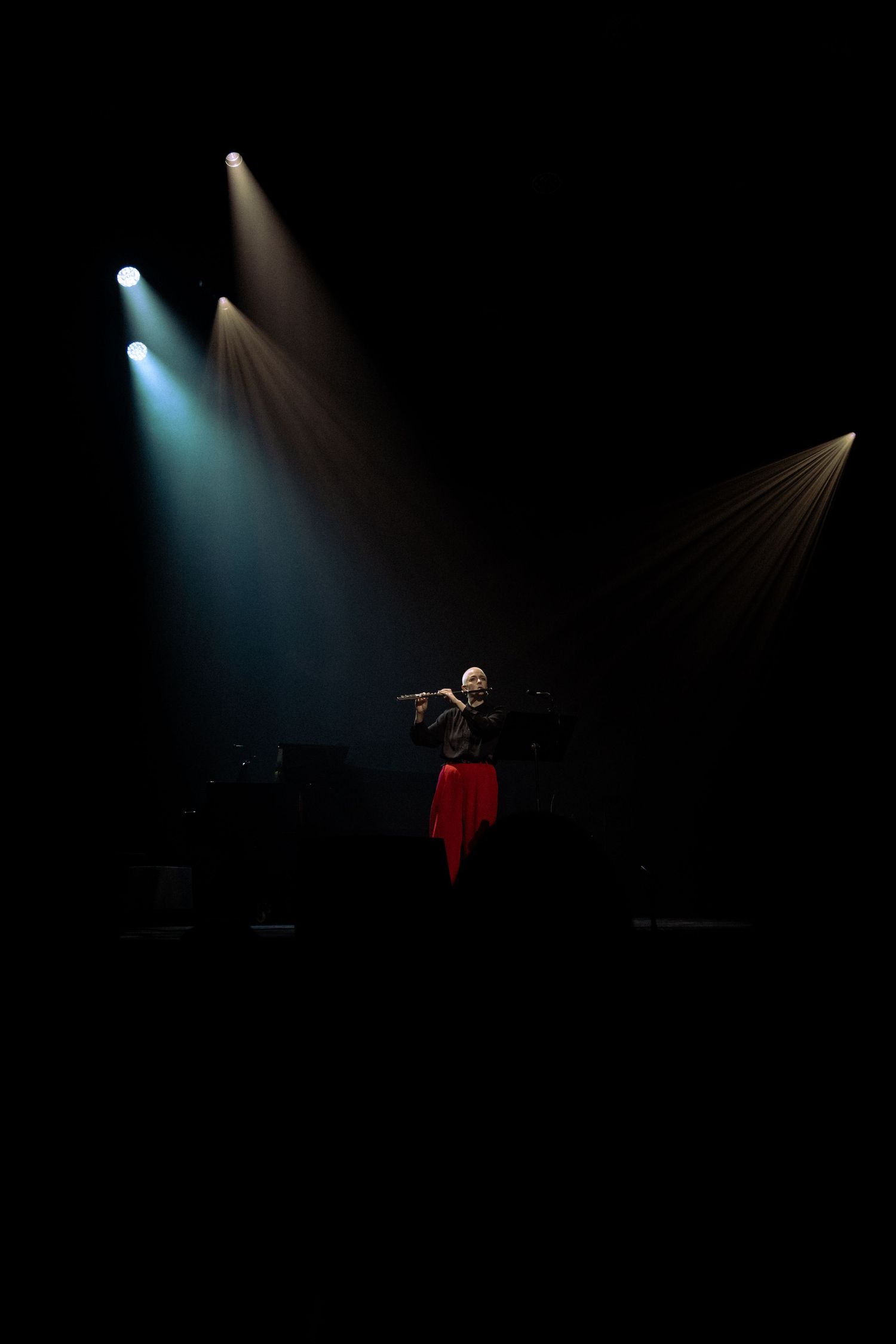 A flutist in a red skirt performs on a dark stage illuminated by dramatic spotlights during a live concert.