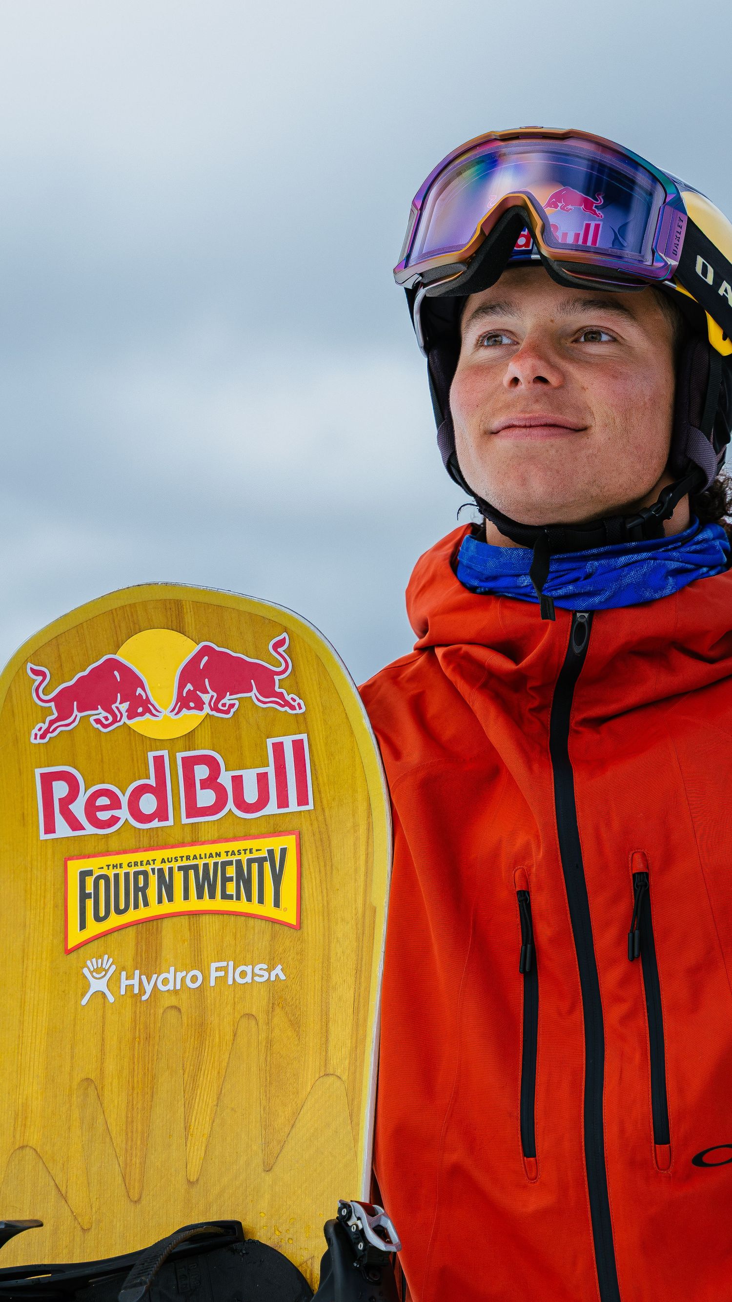 Young snowboarder holding a Red Bull, Four'N Twenty, and Hydro Flask branded yellow snowboard, wearing an orange jacket and goggles.