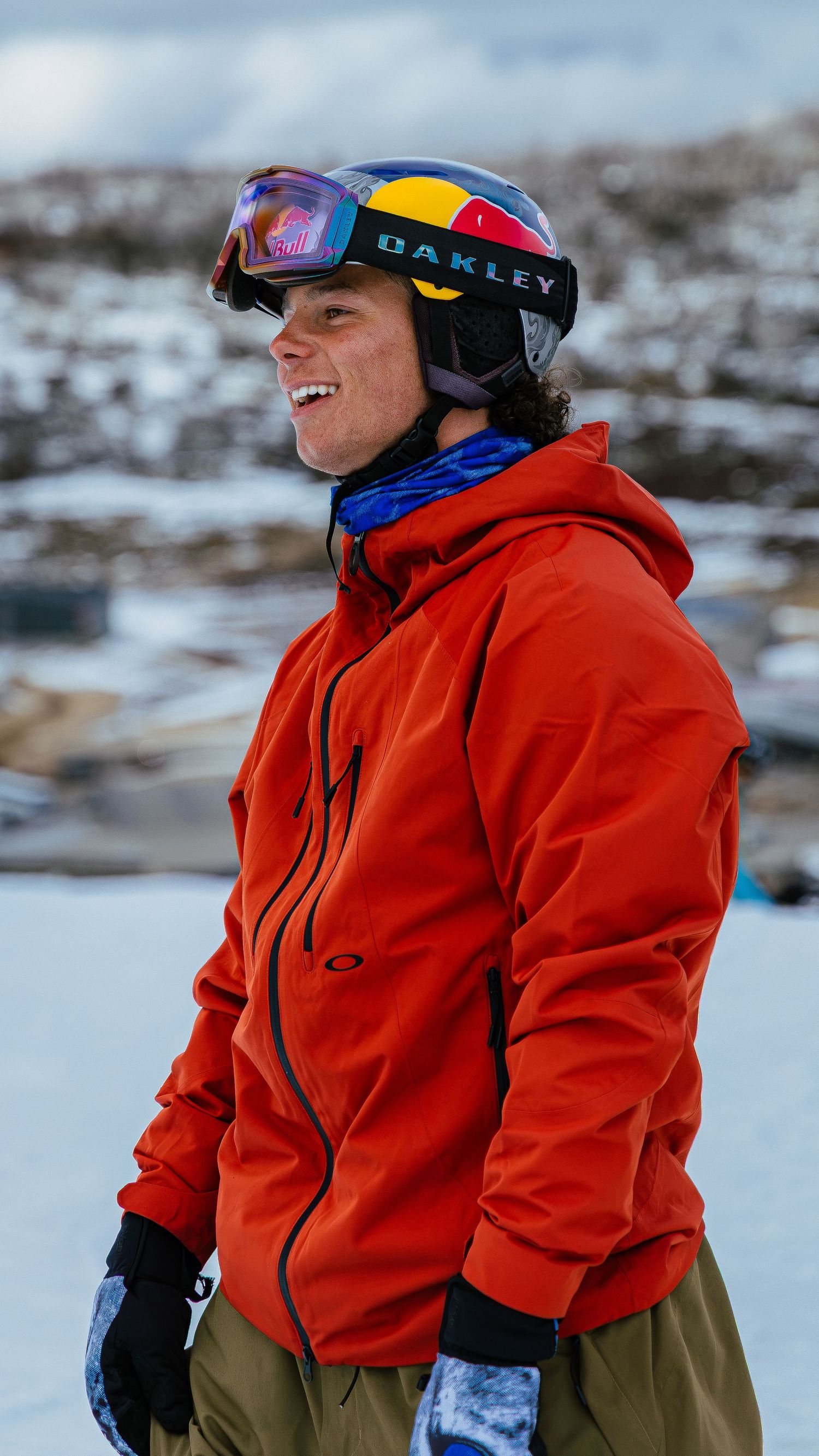 Smiling snowboarder wearing red Oakley jacket and helmet with Red Bull goggles standing on snowy mountain slope.