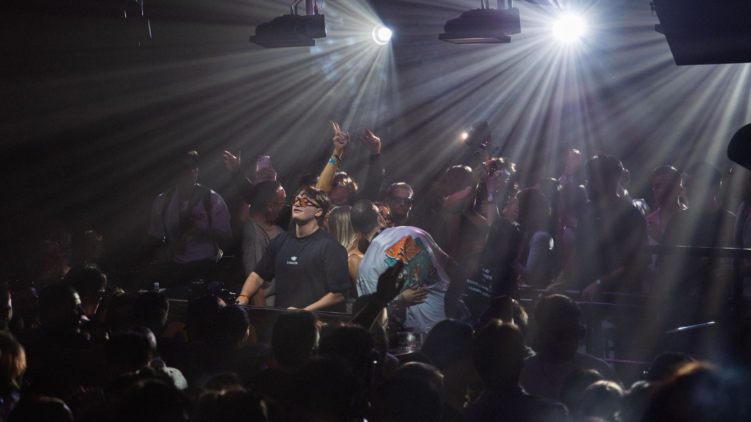 Crowd at a nightclub concert with dramatic stage lighting beams illuminating performers and fans.