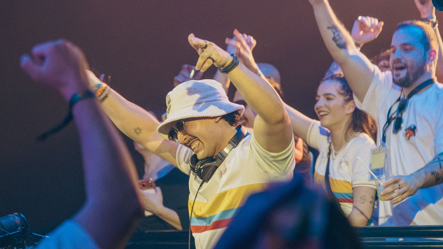 DJ in bucket hat with headphones raises hands at lively music festival crowd cheering behind him.