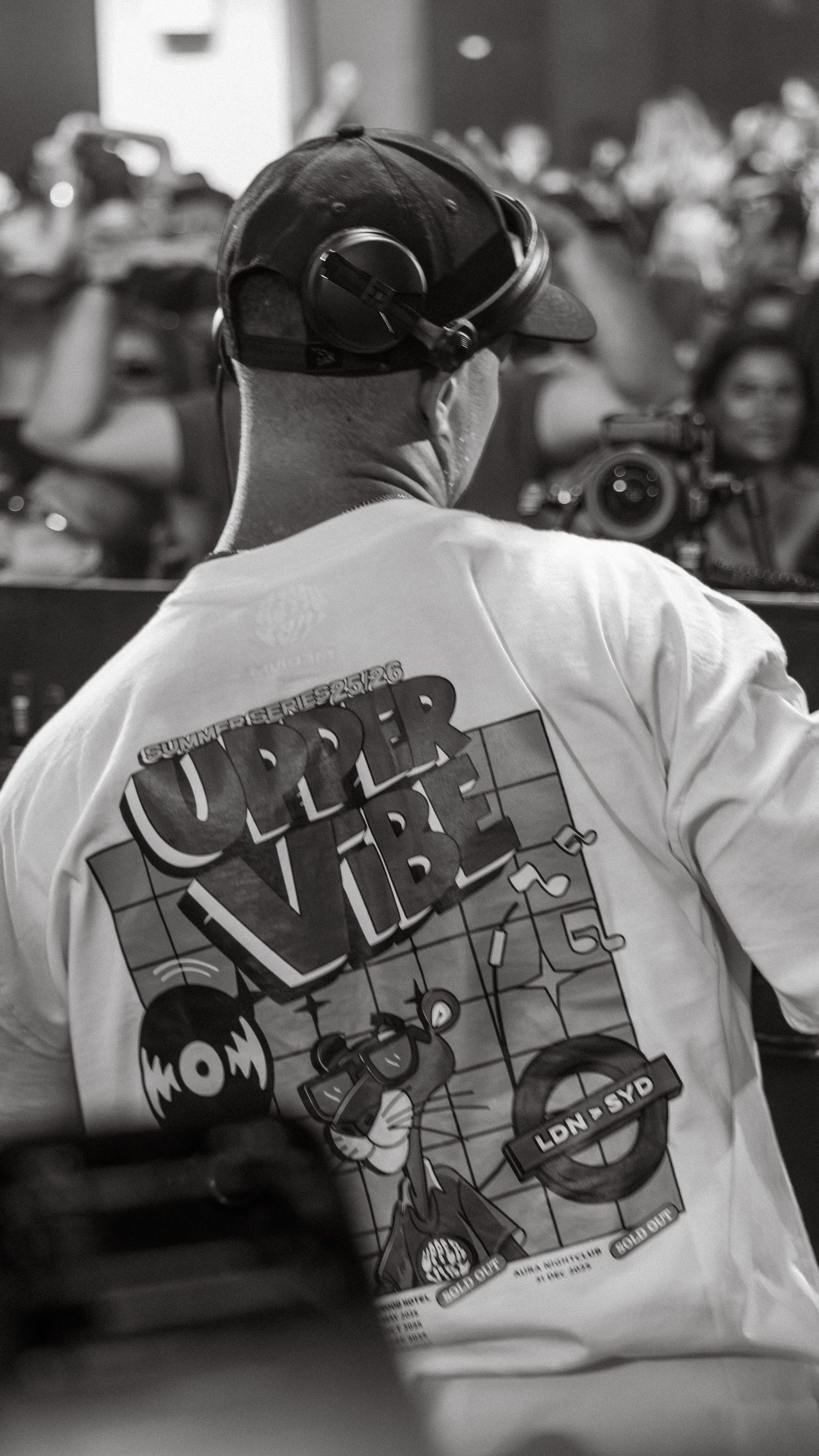 Black and white photo of a DJ wearing a white Upper Vibe graphic tee and cap, performing at a crowded music event.