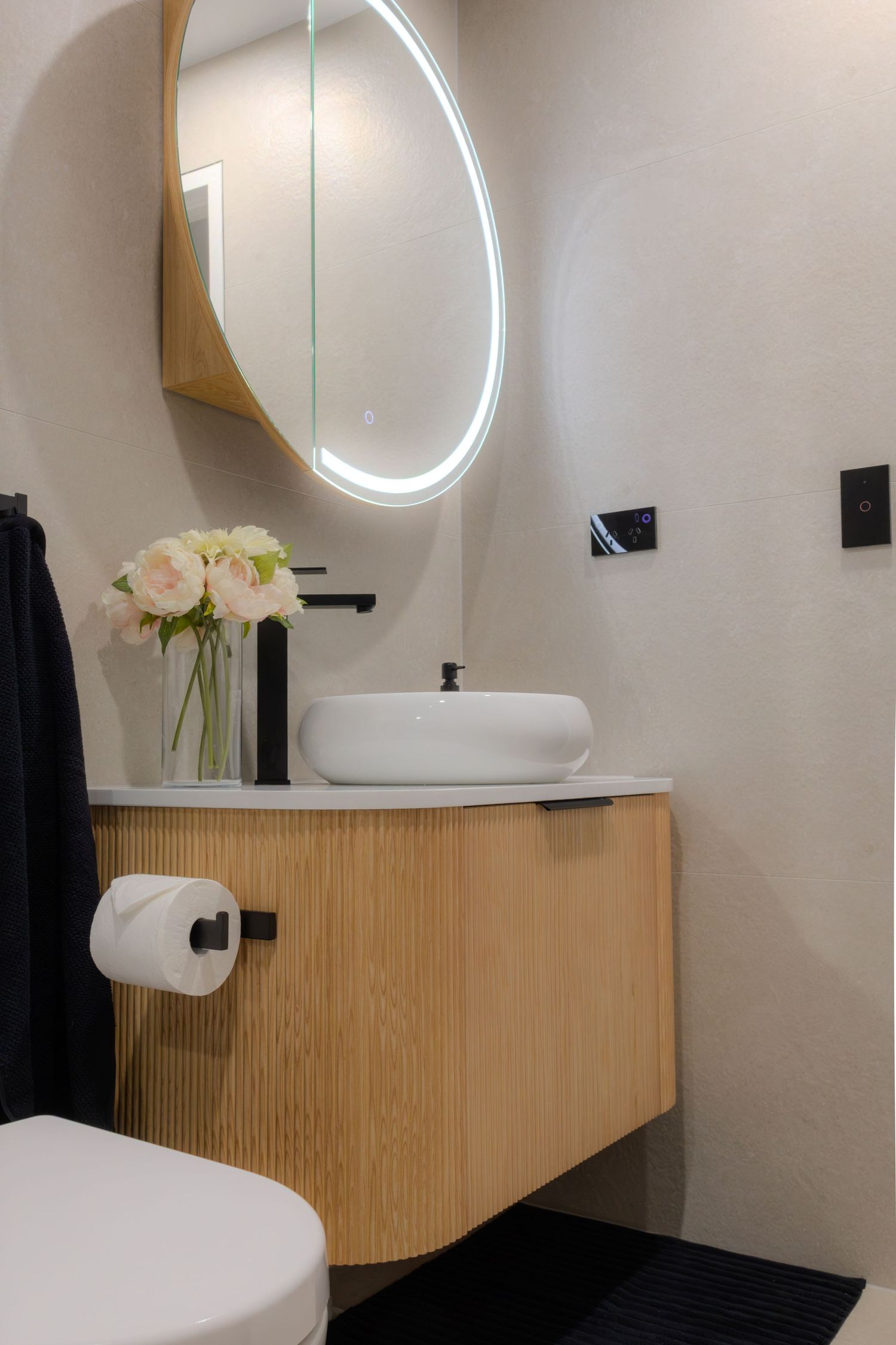 Modern bathroom vanity with wooden cabinet, vessel sink, black faucet, round LED mirror, and fresh flowers.