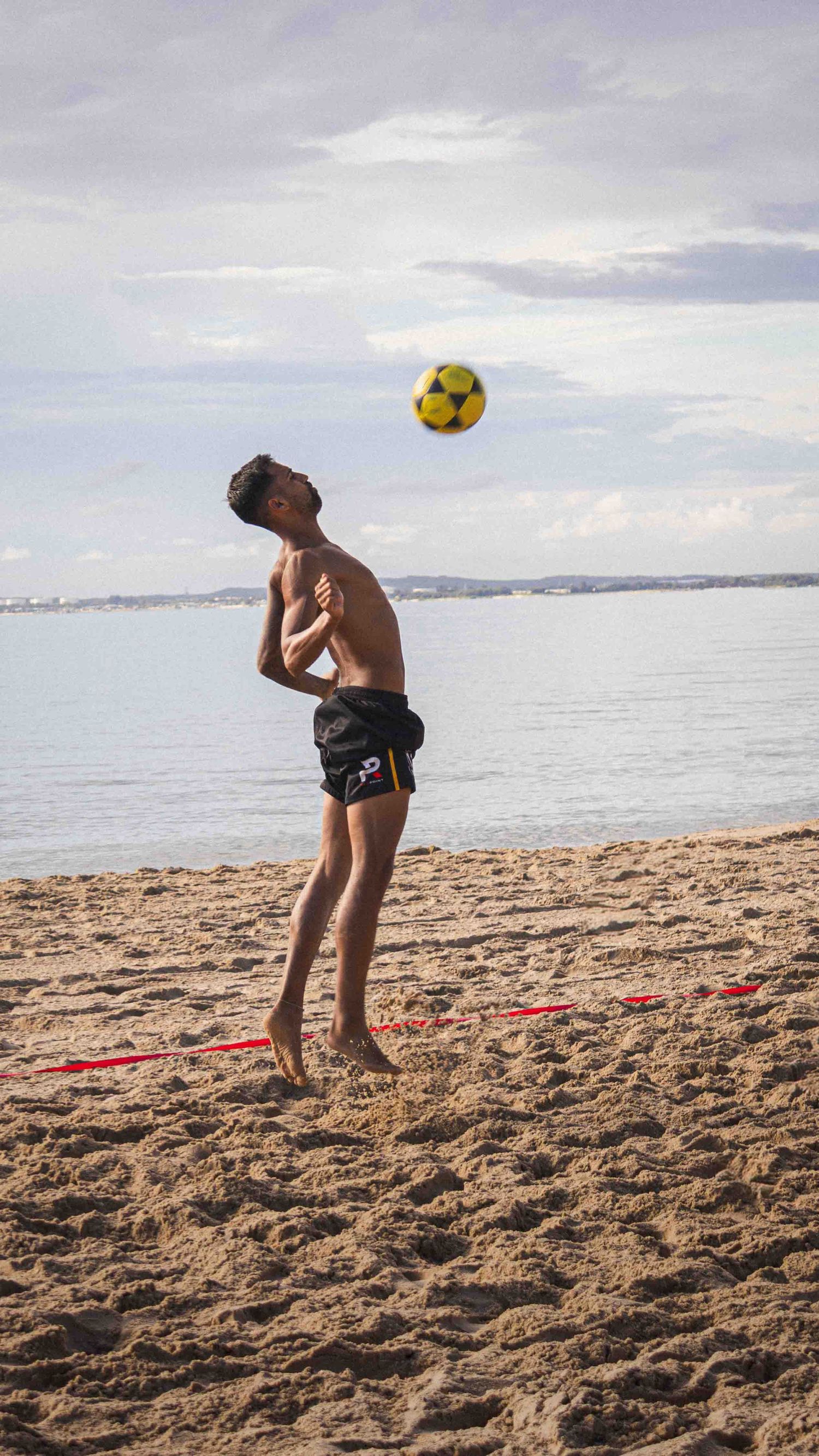 A person practicing soccer ball tricks on a sandy beach with ocean and cloudy sky in the background.