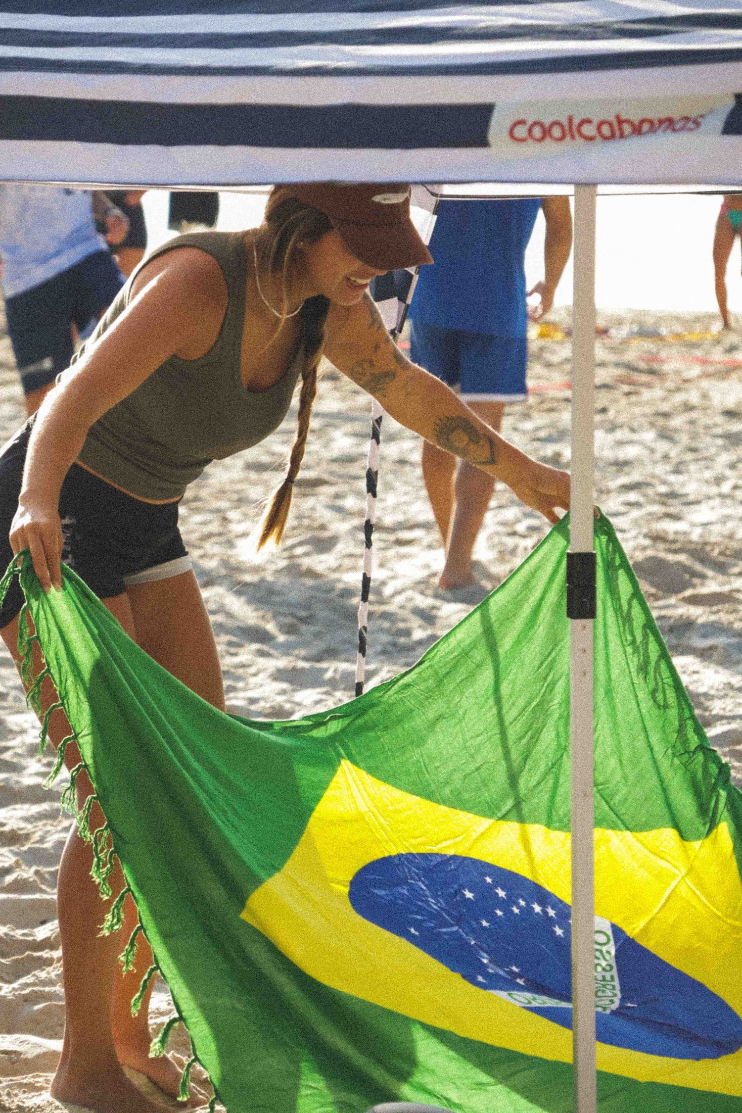 Woman holding Brazilian flag on sandy beach under a coolcabanas tent in bright sunlight.