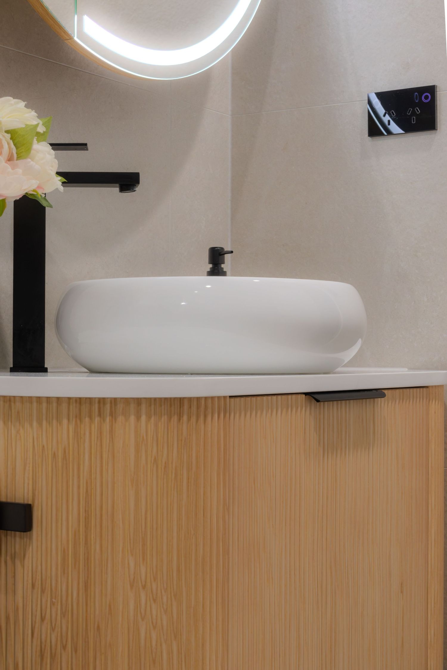 Modern bathroom vanity with white vessel sink, black matte faucet, wood cabinet, and illuminated round mirror.