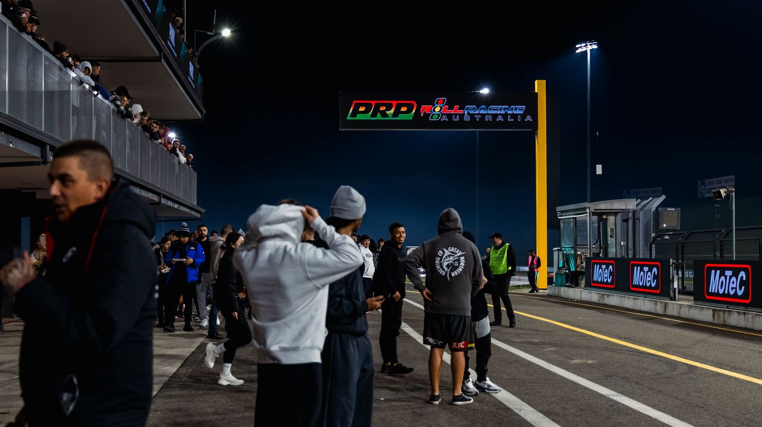 Night scene at PRD motorsport event with spectators and crew members gathered in the pit lane under colorful signage.