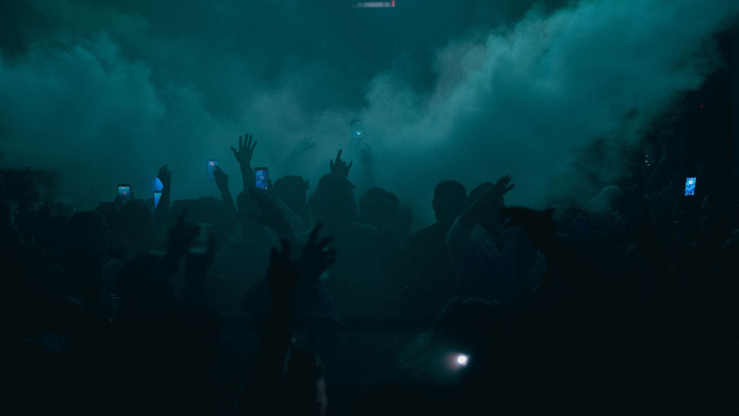 Crowd of silhouetted concert-goers with raised hands in teal-lit smoky atmosphere at a live music event.