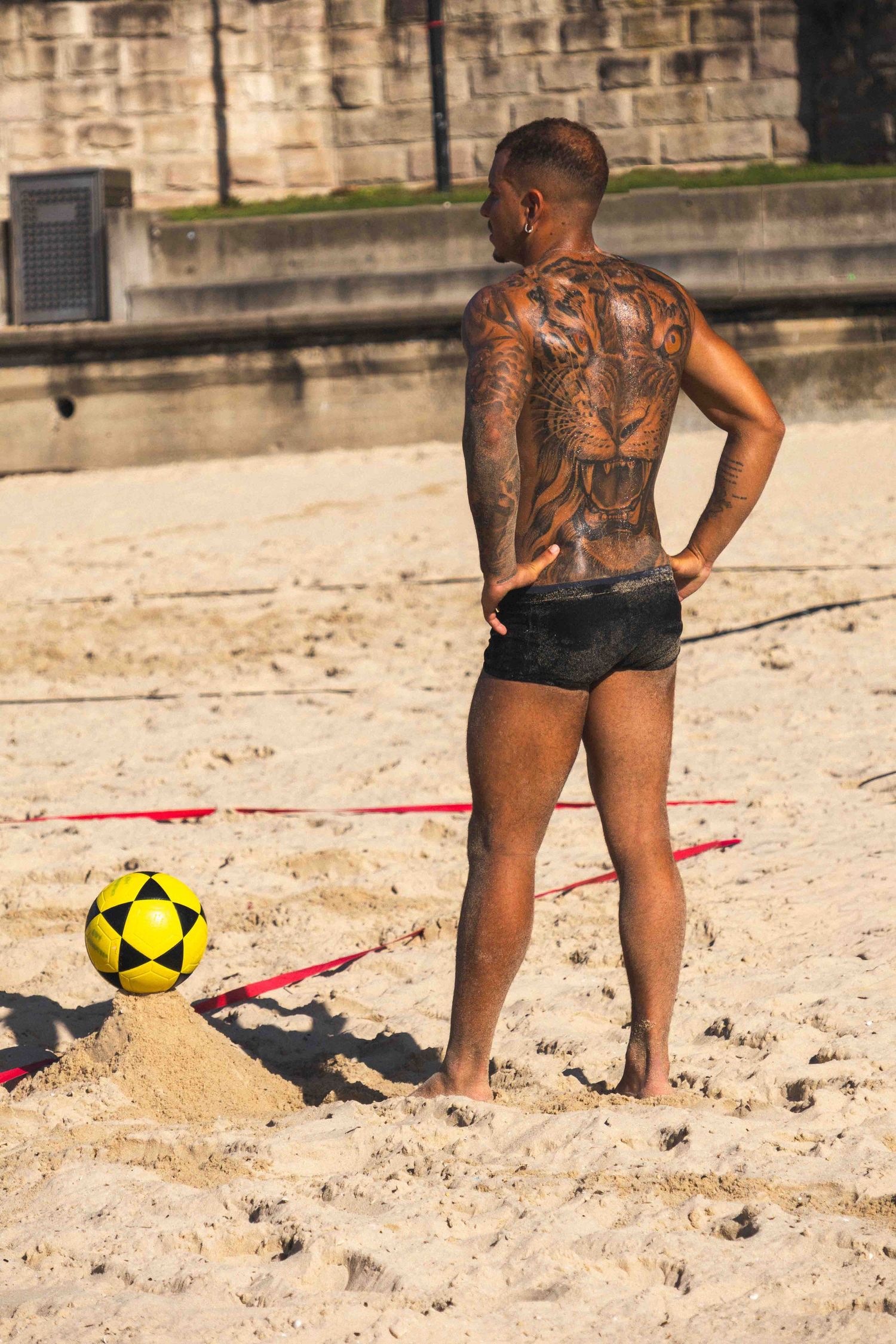Person with elaborate back tattoos plays beach volleyball near a yellow ball on sandy terrain.