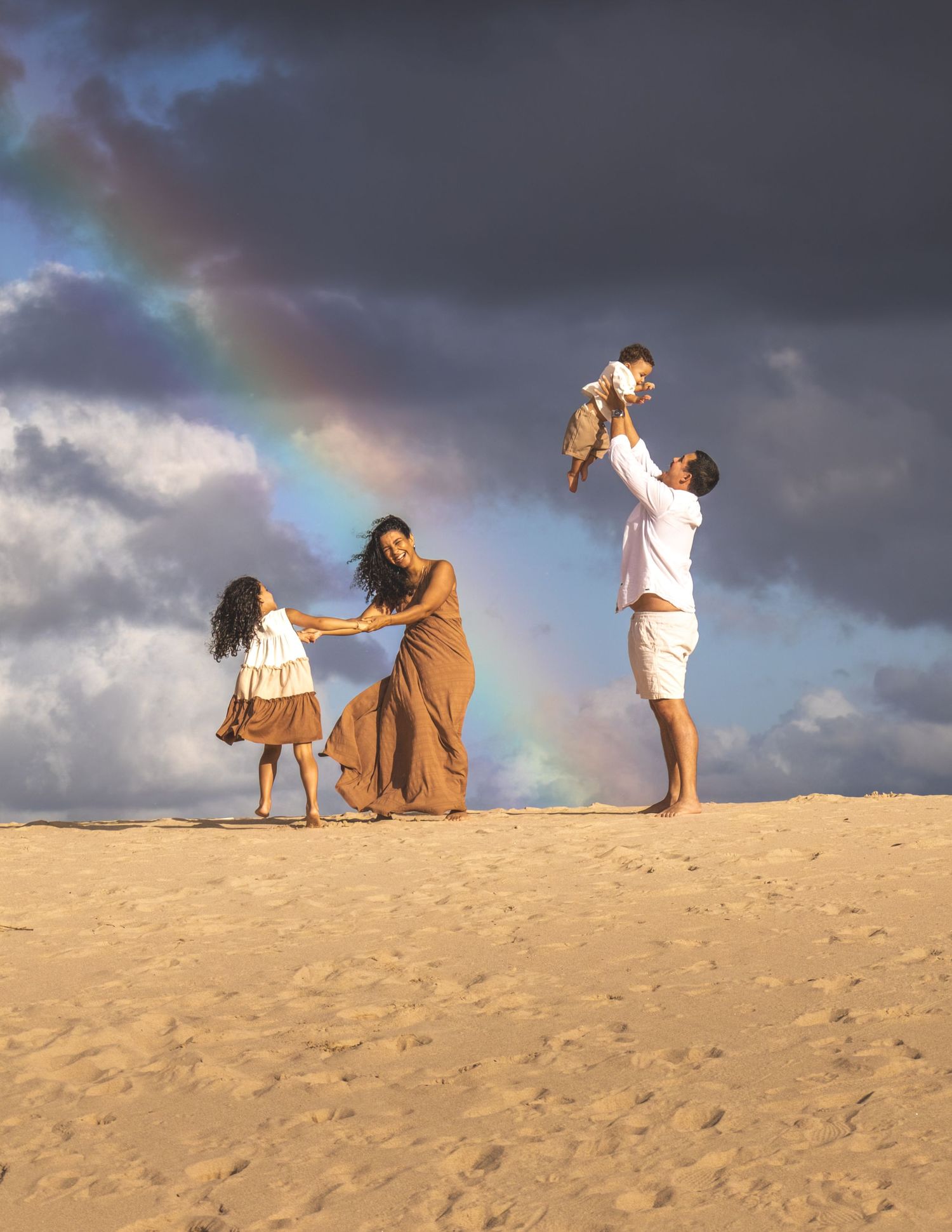 Family plays on sandy dunes at sunset with dramatic clouds and rainbow in sky.
