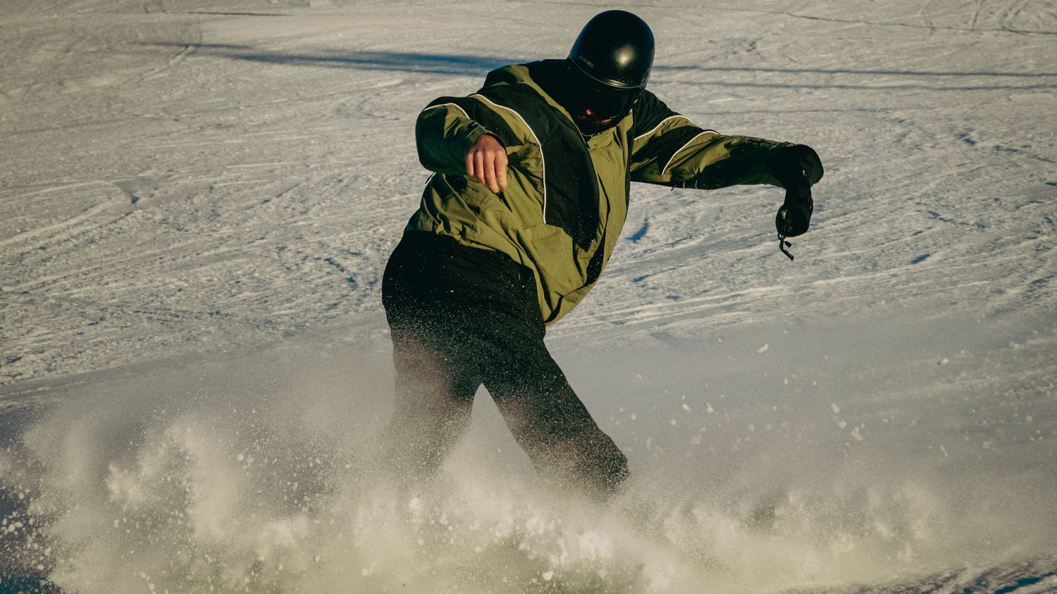 Snowboarder carving through fresh powder snow on a mountain slope wearing a green jacket and black helmet.