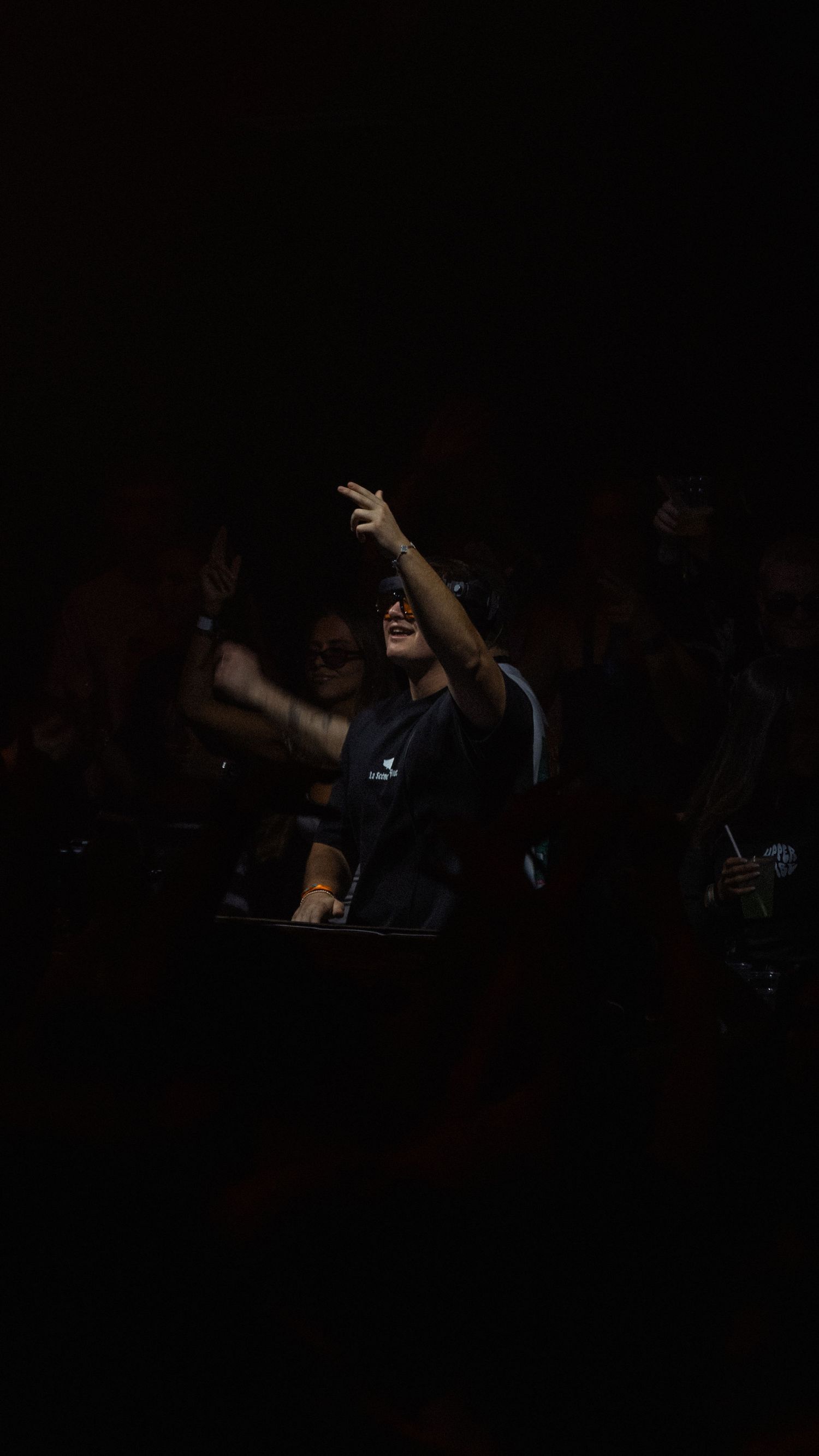 A DJ performs energetically in a dark venue, raising their hand while standing behind equipment at a nightclub event.