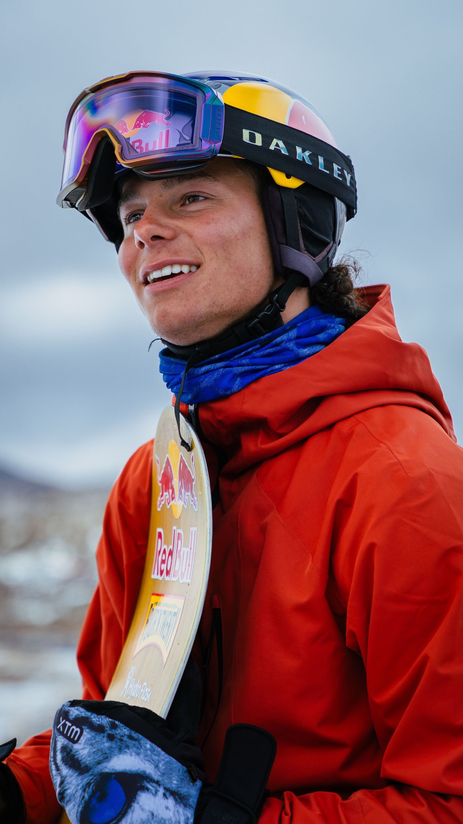 Young male snowboarder smiling in Oakley goggles, Red Bull helmet, and orange jacket holding a snowboard outdoors.