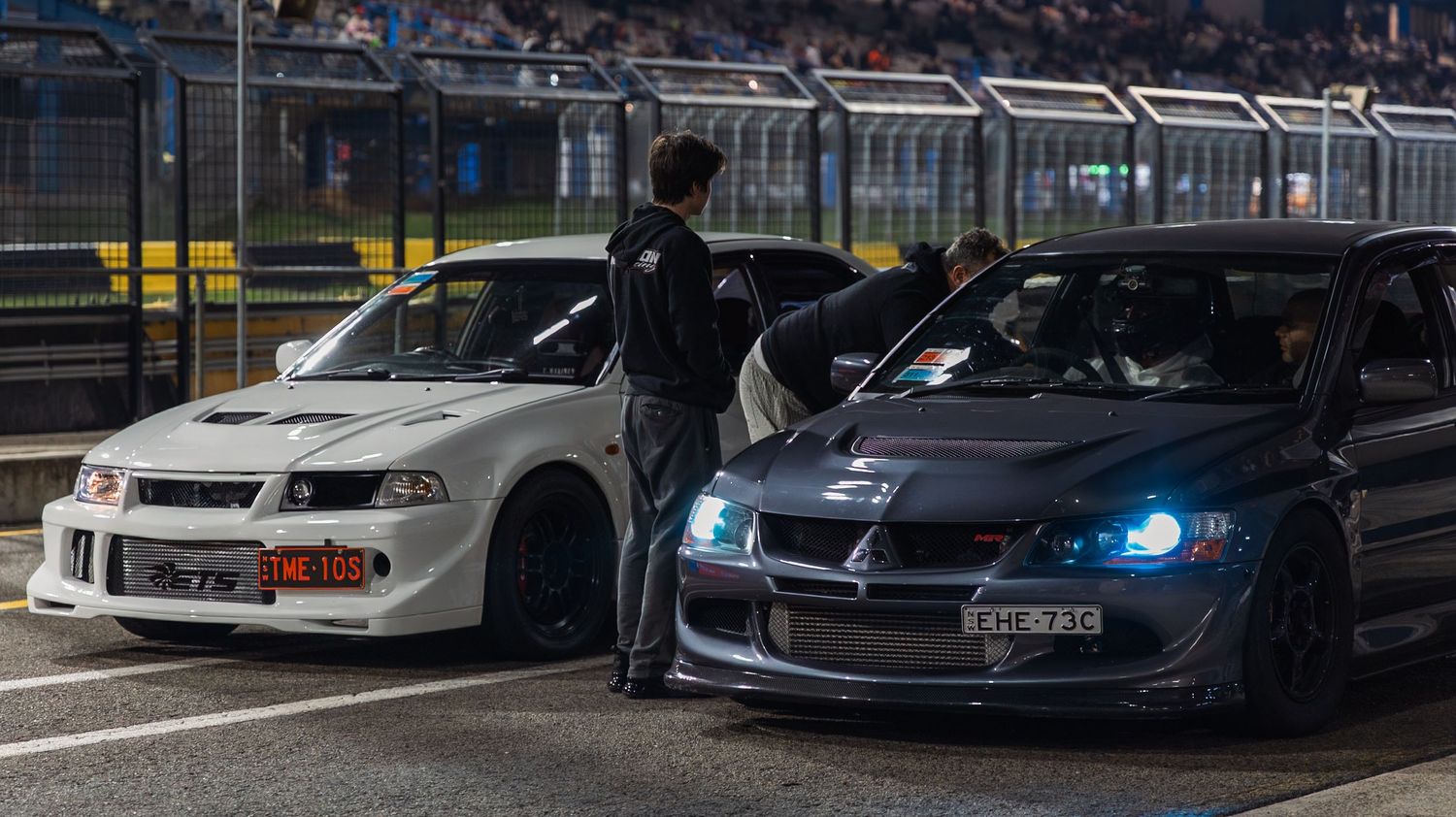 Two modified Mitsubishi Lancer Evolution cars parked at a night racing event with blue LED lights and a person standing between them.
