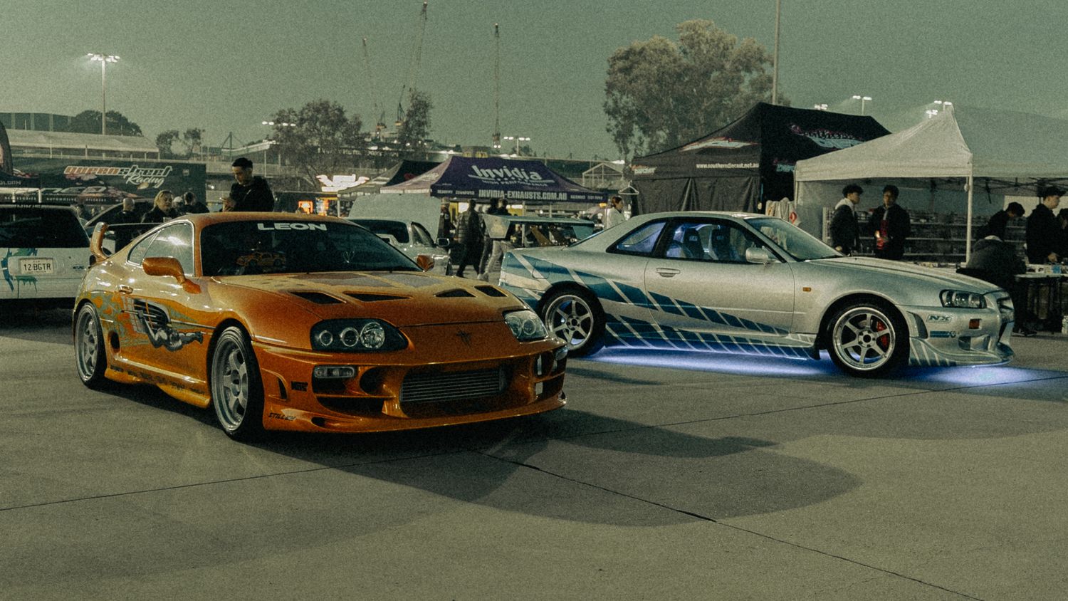 Orange Toyota Supra and silver Nissan Skyline GT-R from Fast and Furious parked at an outdoor car show.