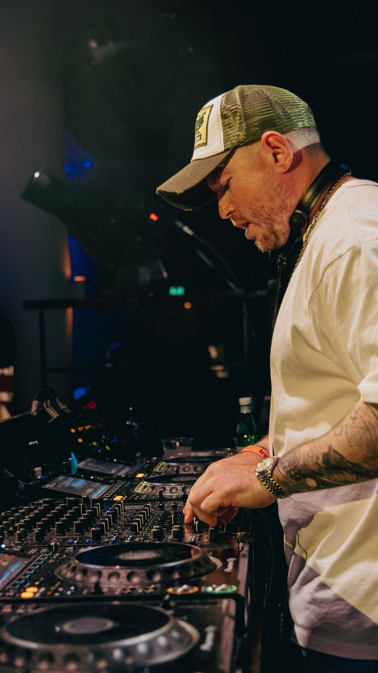 Tattooed DJ wearing a trucker hat and white tee performs at a nightclub mixing board under dramatic stage lighting.