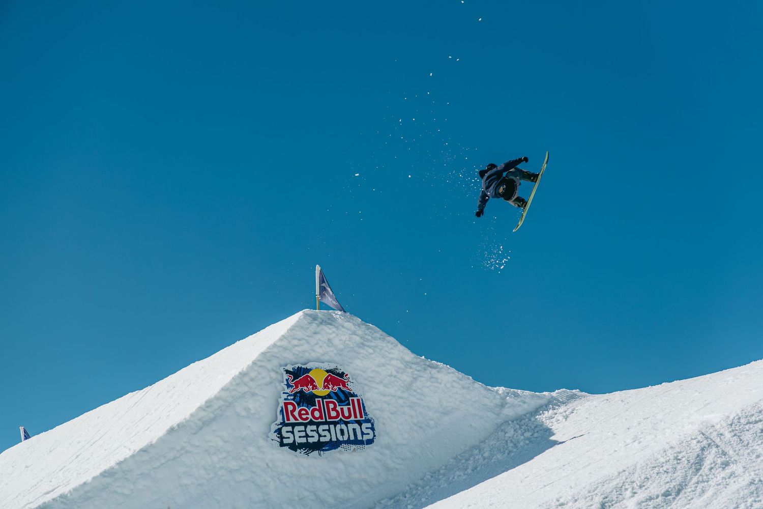 Snowboarder performs high aerial trick over Red Bull Sessions branded snow ramp against clear blue sky.
