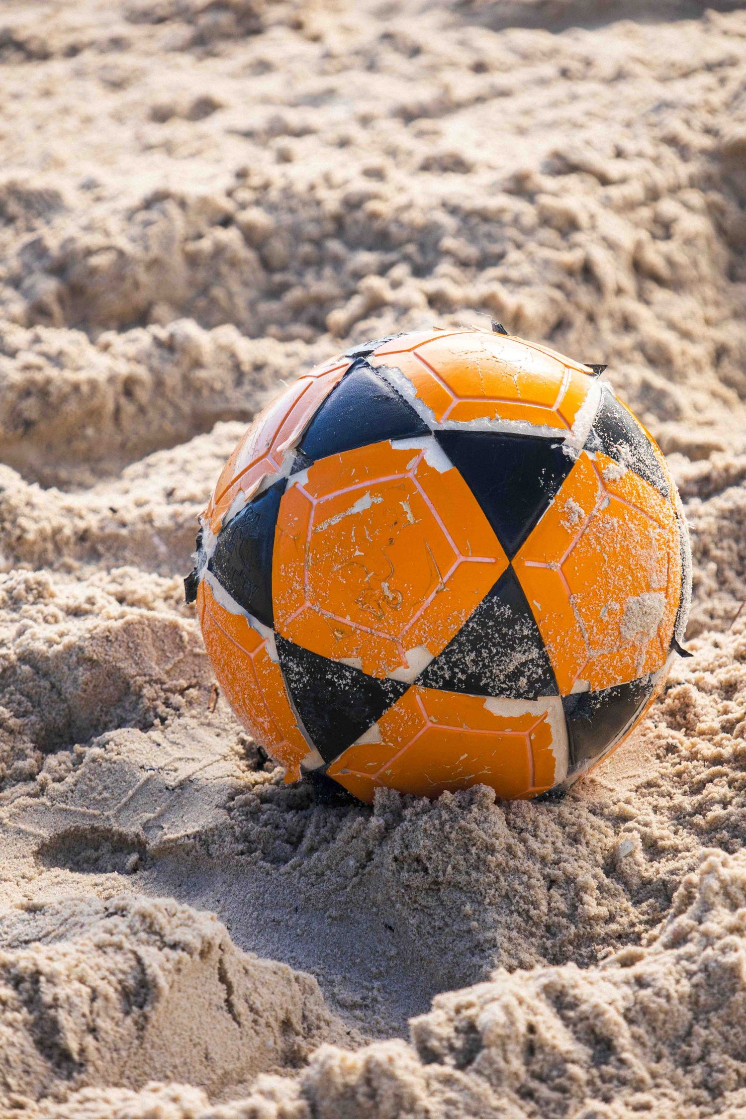 An orange and black soccer ball sits partially buried in sandy beach terrain on a sunny day.