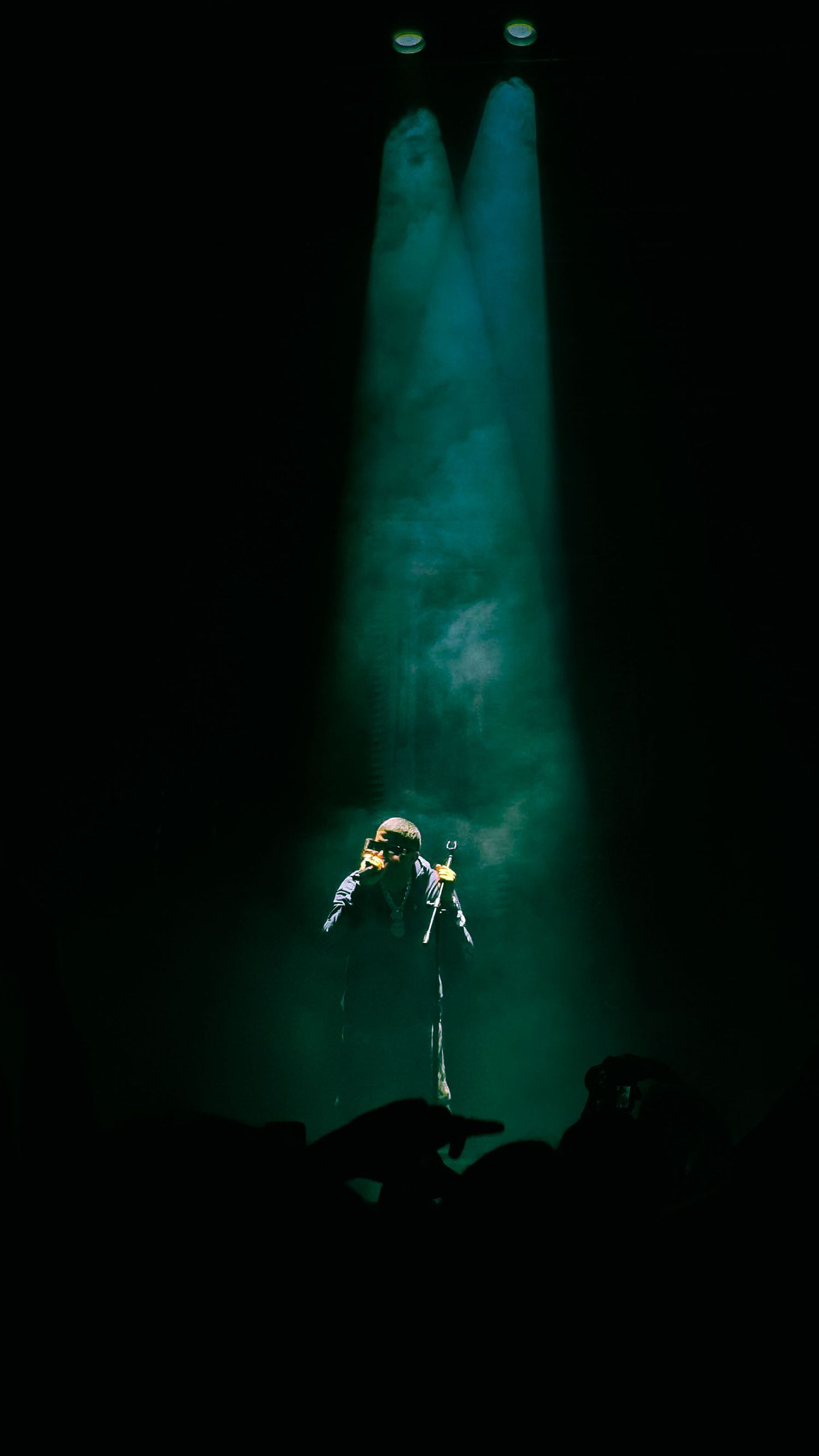 A performer stands alone on a dark stage, illuminated by dramatic green spotlights with atmospheric smoke swirling above.