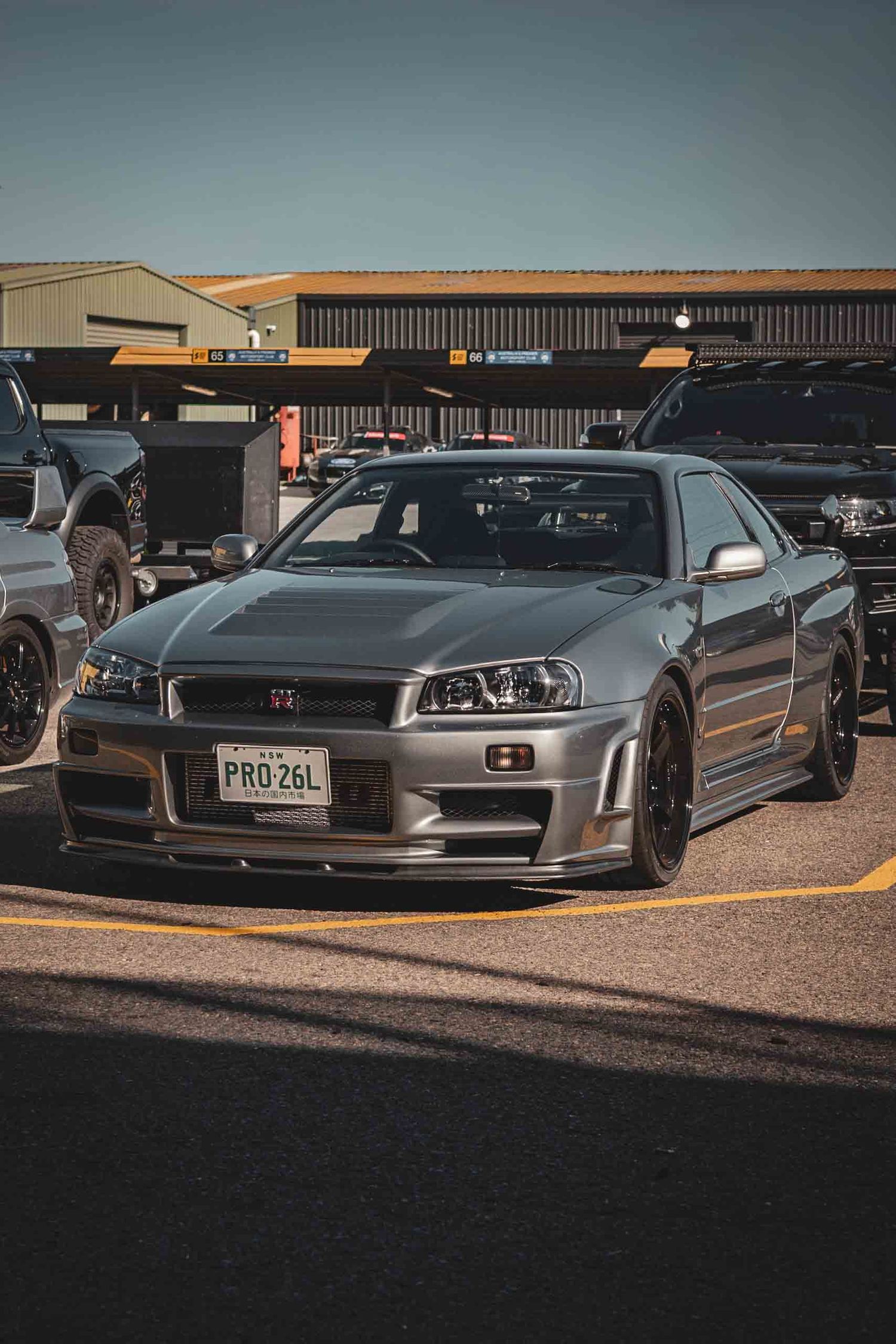 Silver Nissan Skyline R34 GT-R with license plate PRO 26L parked in a lot, featuring black wheels and aggressive styling.