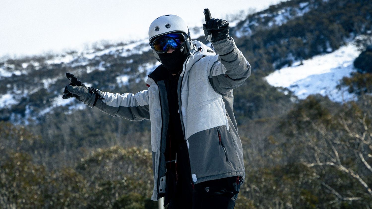 Snowboarder in white helmet and goggles giving thumbs up on snowy mountain slope.
