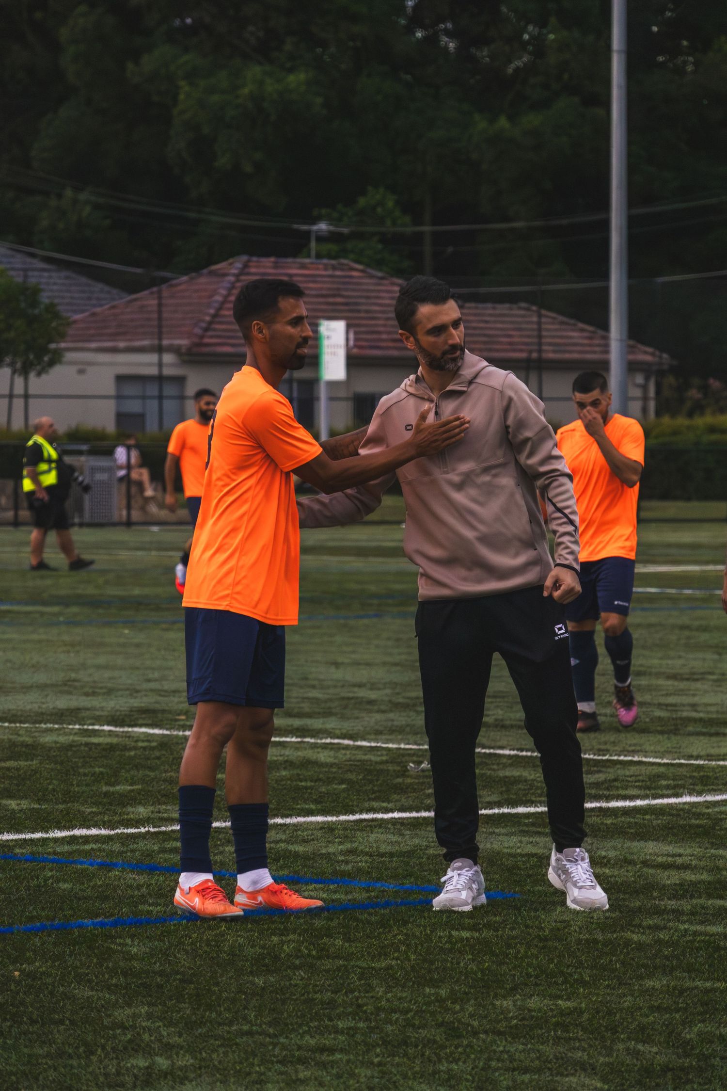 Soccer coaches demonstrate training techniques on a grass field while wearing orange and tan colored shirts.