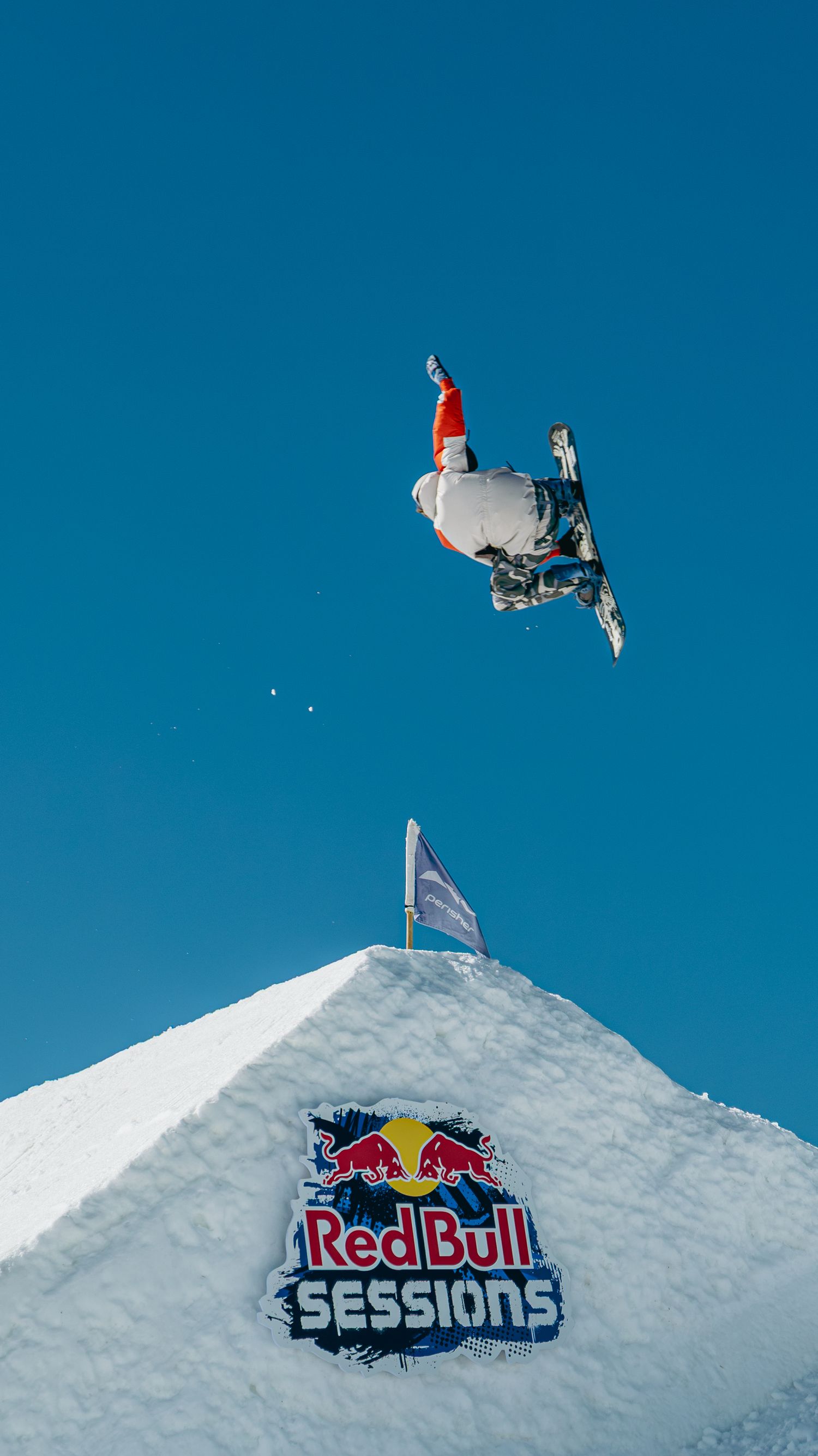Snowboarder performs aerial trick above a snow ramp at Red Bull Sessions event under clear blue sky.