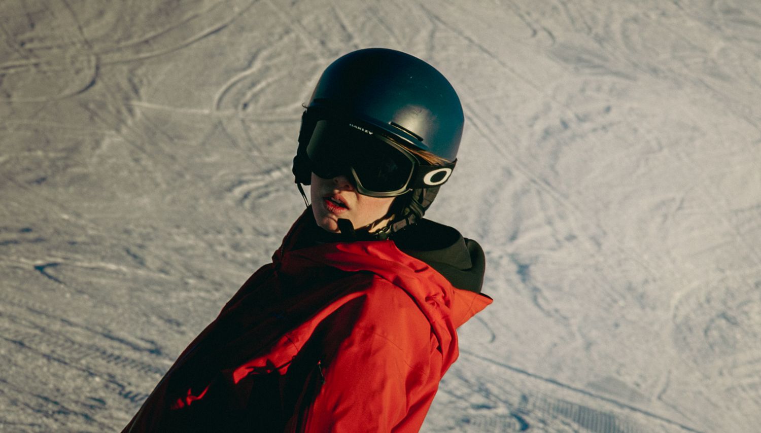 A snowboarder in a red jacket and Oakley goggles with a dark helmet poses on a snowy mountain slope.