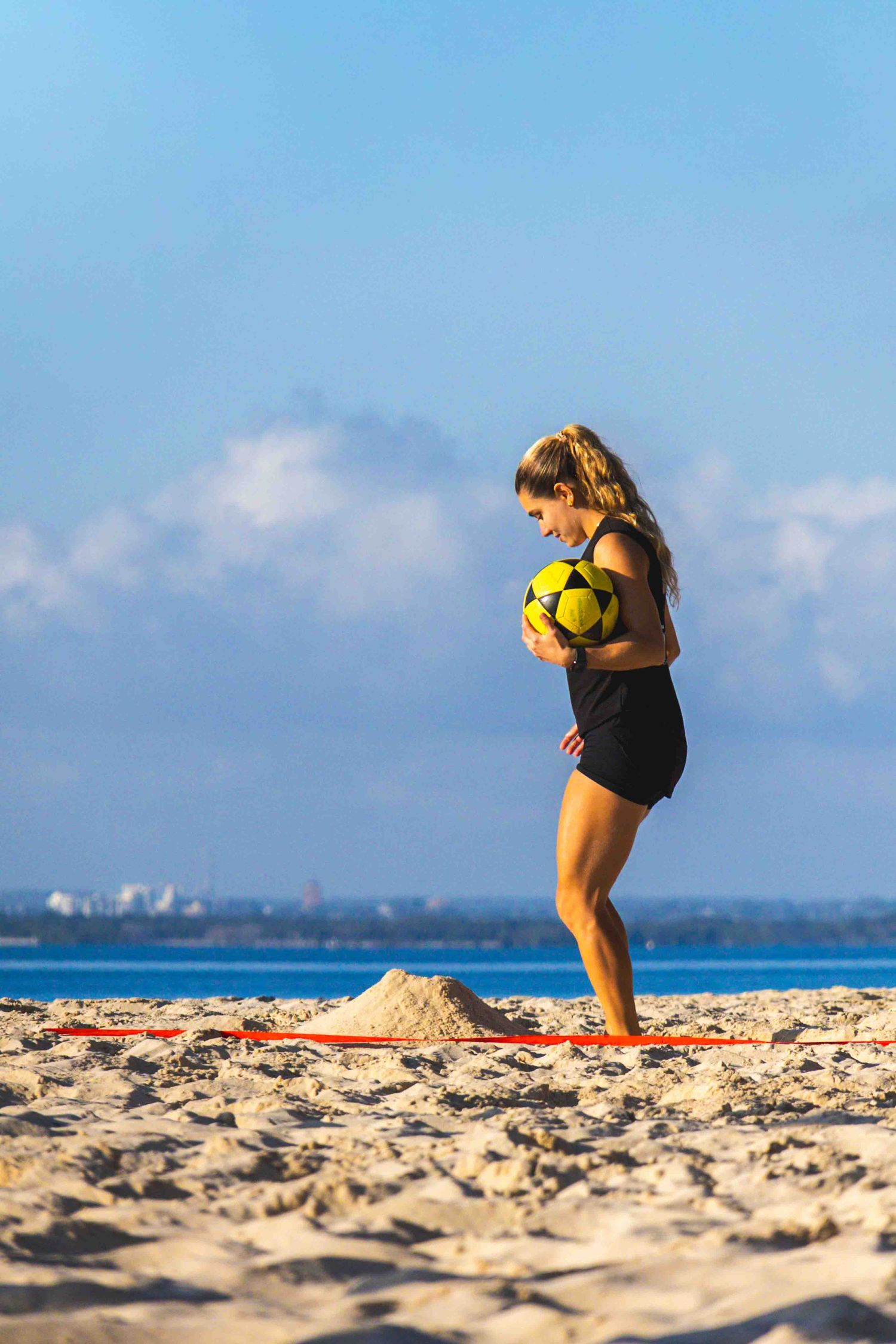 Woman in black swimsuit balancing on slackline at beach while holding yellow soccer ball on sunny day.