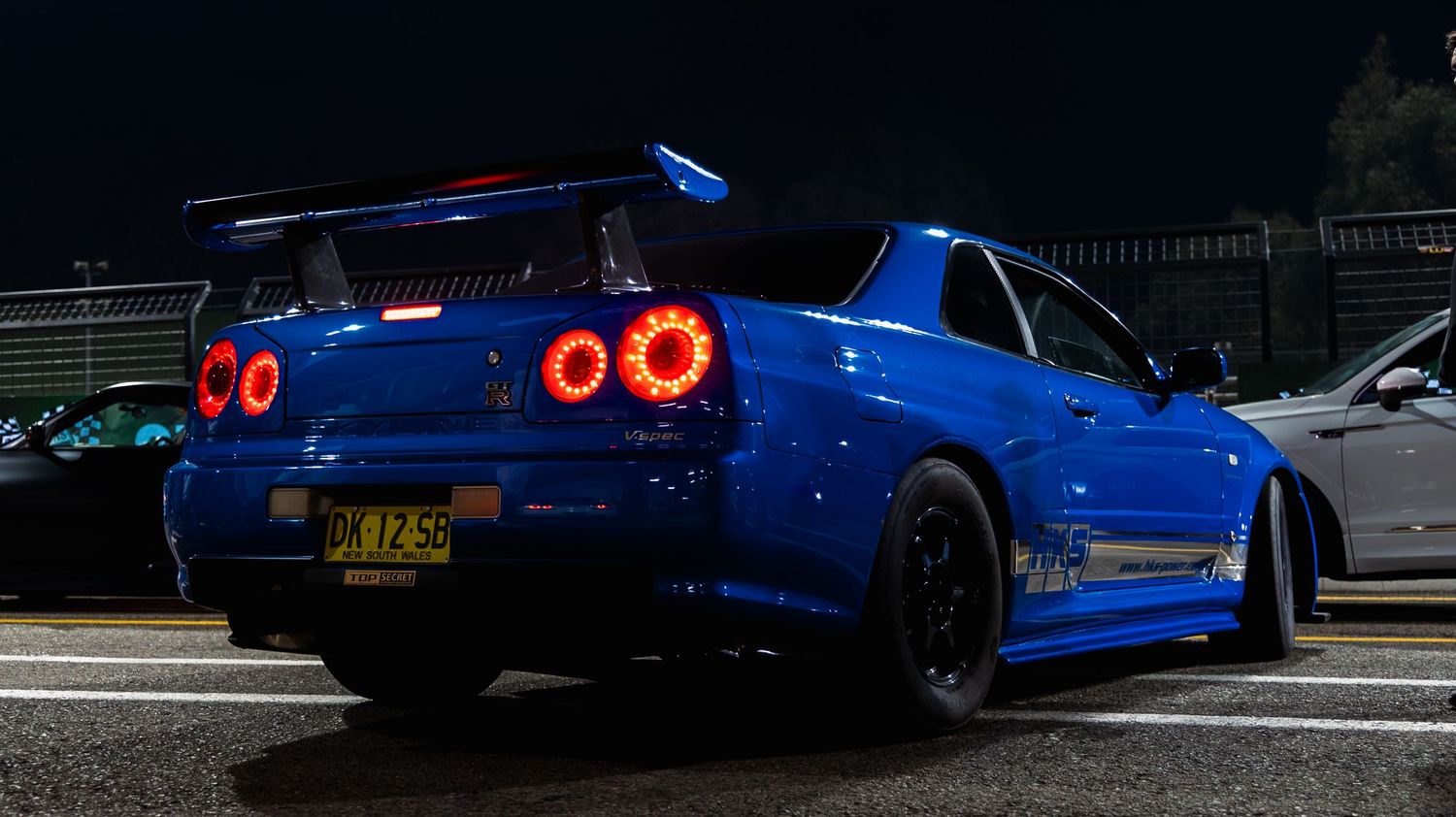 Blue Nissan Skyline R34 GT-R with red halo headlights and rear spoiler photographed at night.